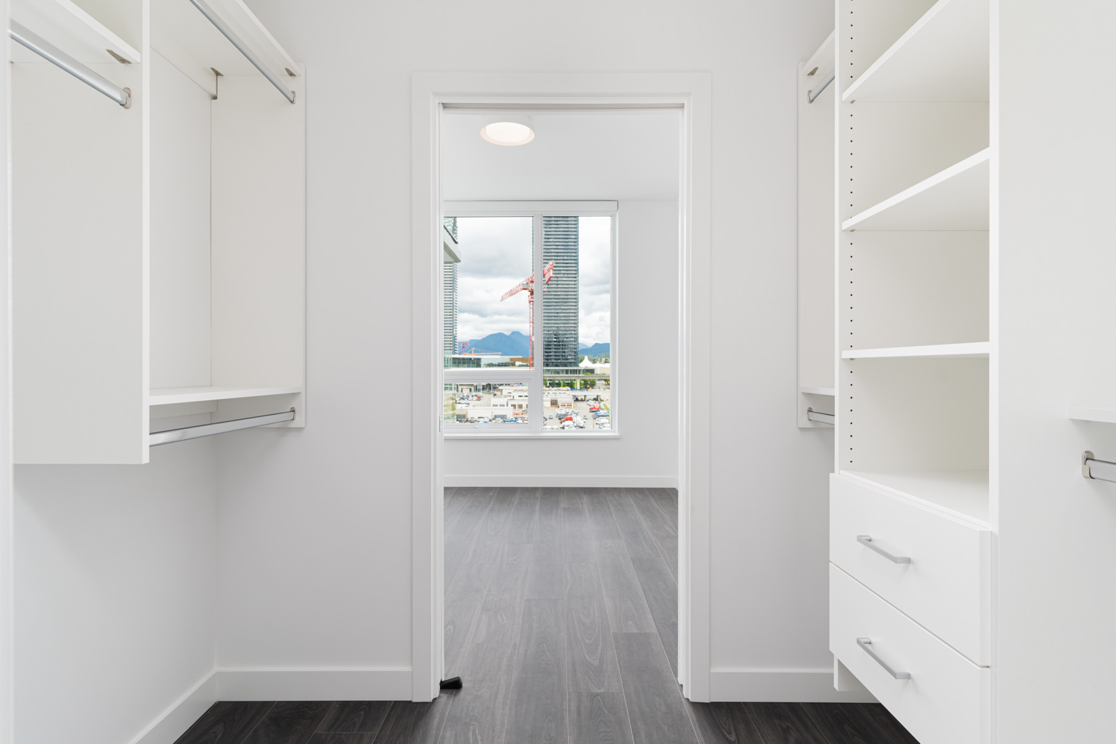 A white walk-in closet with shelves and hanging rods, leading to a bright room with large windows and a cityscape view outside.