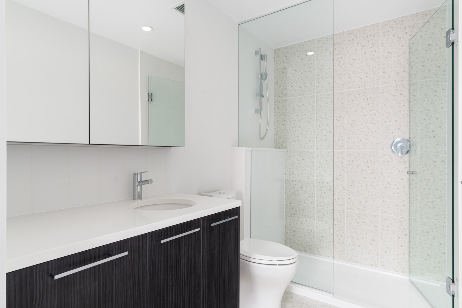 Modern bathroom with a dark wood vanity, white countertop, wall-mounted faucet, toilet, and a glass-enclosed shower with light beige tile.