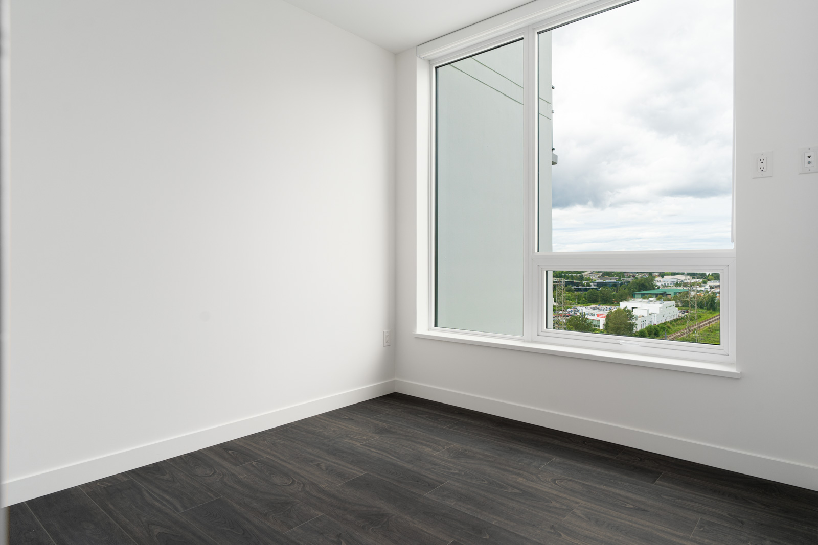 Empty room with white walls, dark wood flooring, and a large window showing an outdoor view with buildings, greenery, and a cloudy sky.