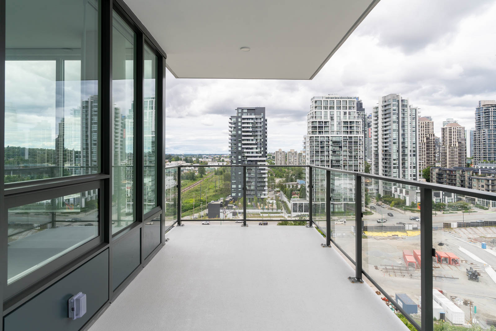 A modern high-rise apartment balcony with glass railings overlooks a cityscape of tall buildings and cloudy sky.