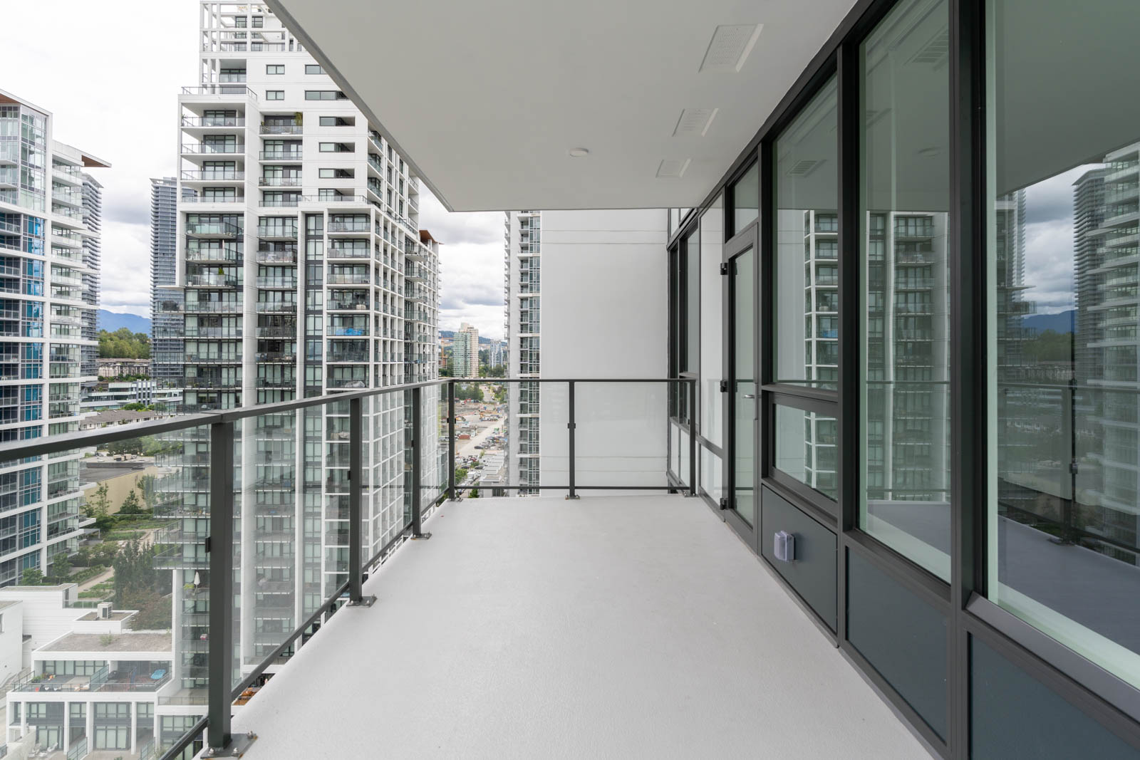 A modern apartment balcony with glass railings, offering a view of nearby high-rise buildings and a cloudy sky.