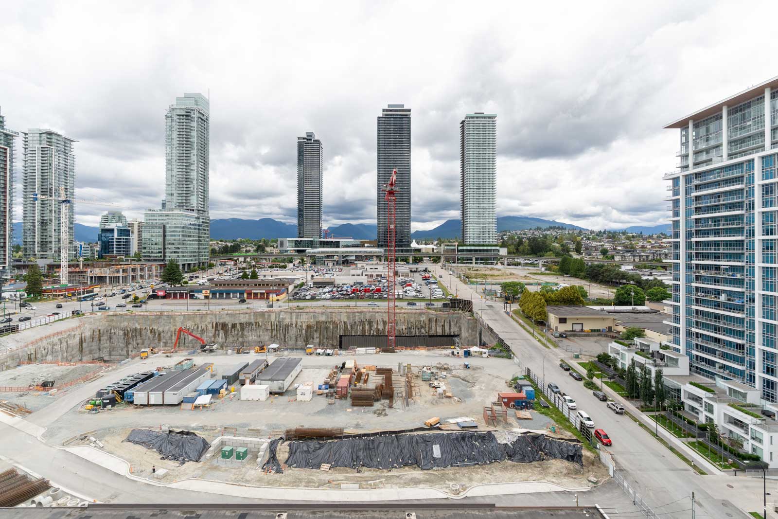 A large construction site with equipment and materials in the foreground, high-rise buildings and cloudy sky in the background, and a view of city streets.