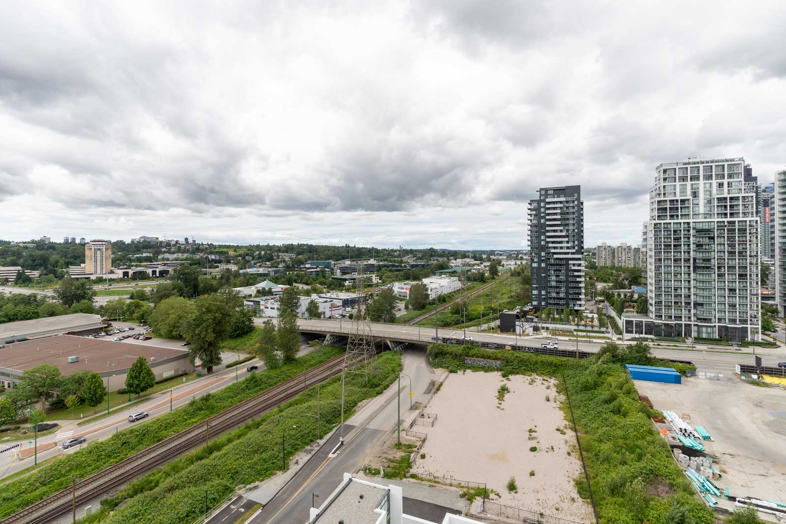 Aerial view of a cityscape with train tracks, roads, modern high-rise buildings, and scattered greenery under a cloudy sky.