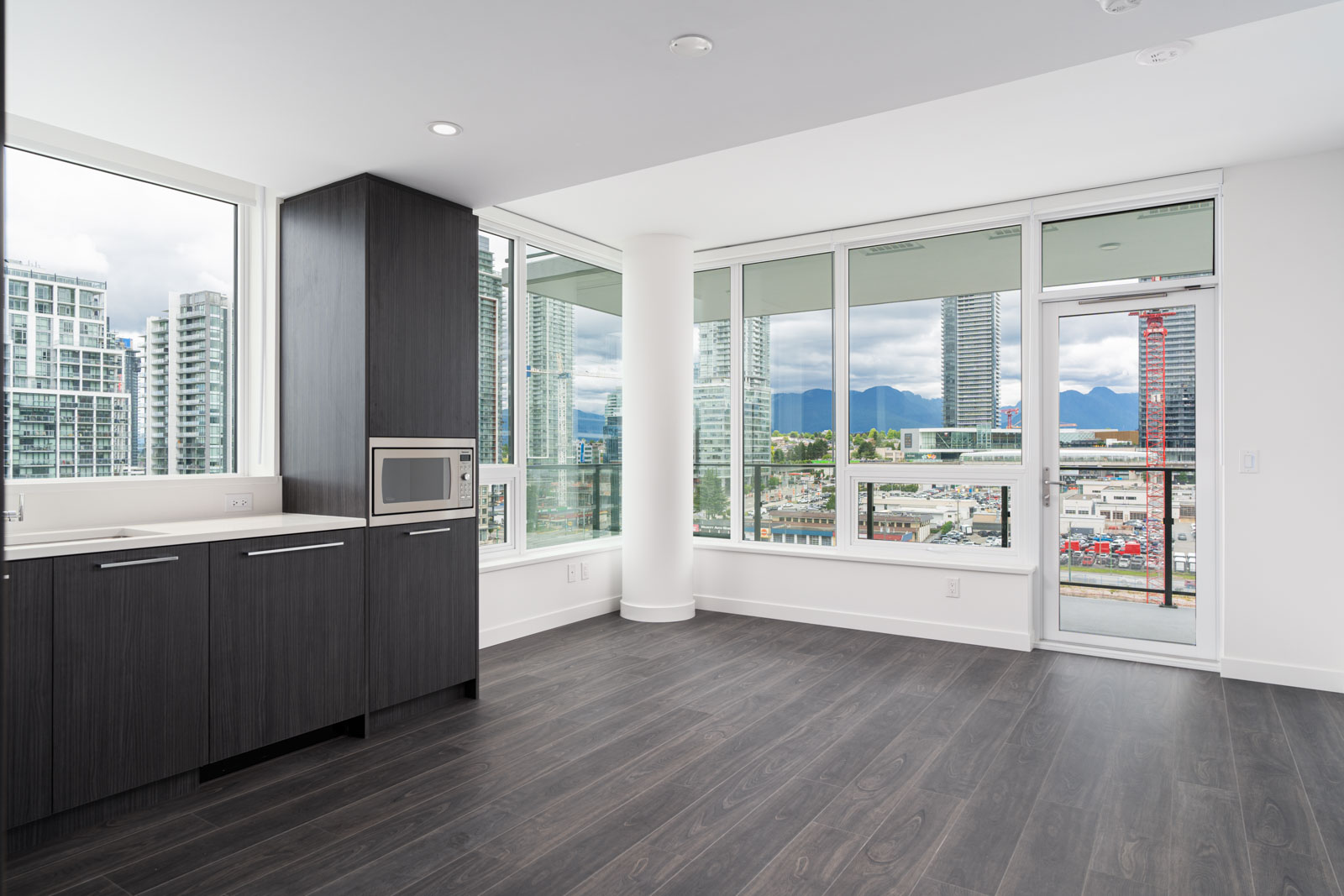 Modern, unfurnished apartment with dark wood floors, large windows, built-in microwave, and balcony overlooking a cityscape with mountains in the background.