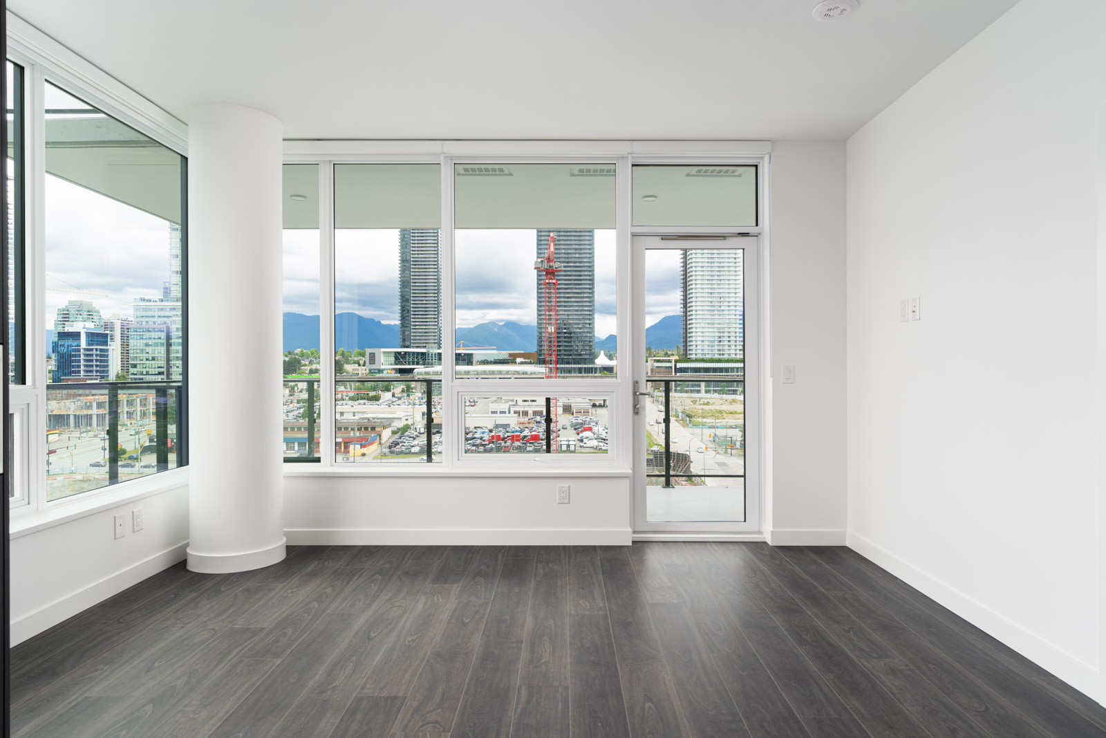 Empty modern apartment room with large windows, glass door to balcony, dark wood floors, and a cityscape view with tall buildings and mountains in the background.