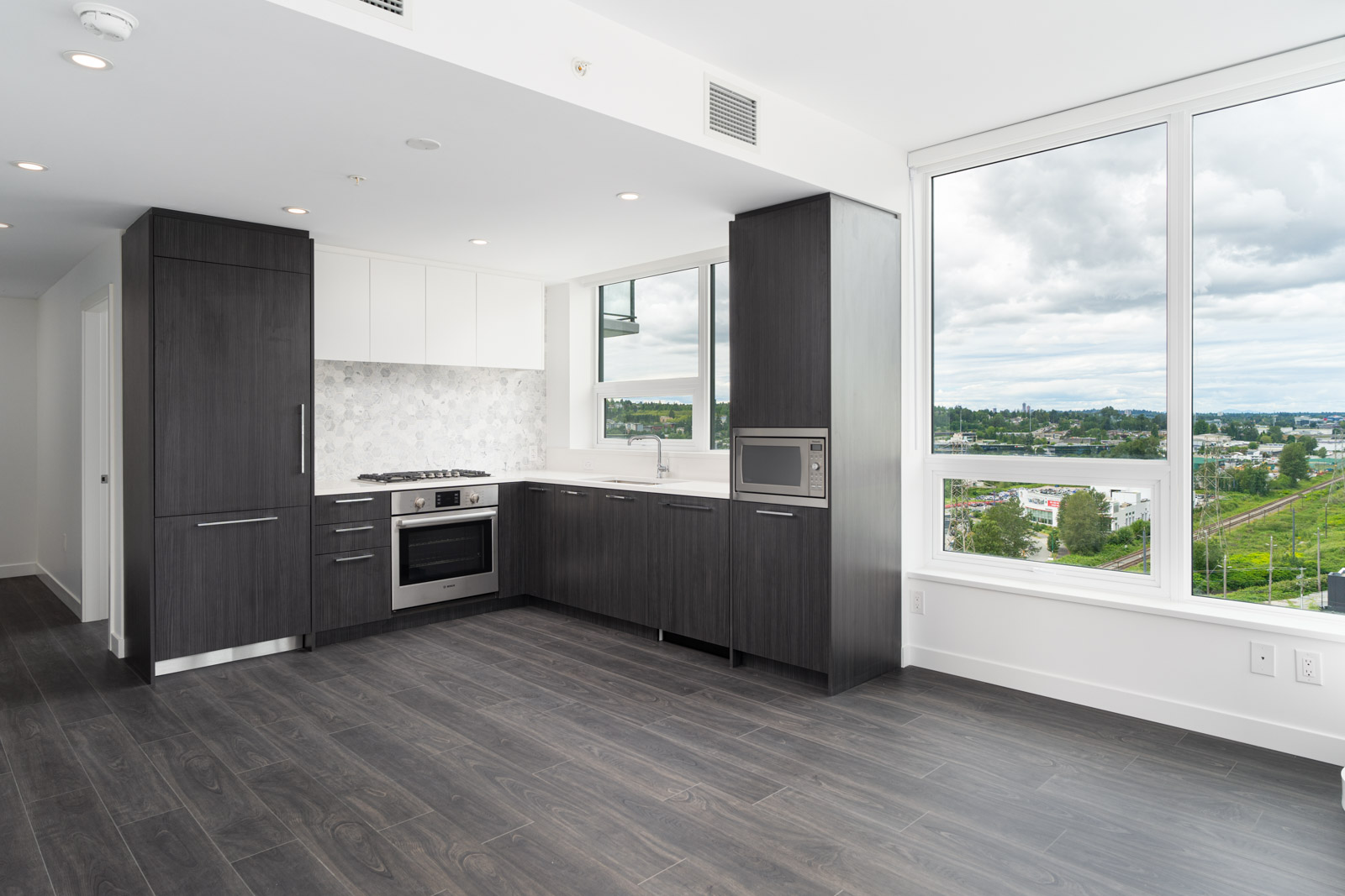 Modern kitchen with dark wood cabinets, built-in appliances, white backsplash, and large windows overlooking a cityscape with greenery and cloudy sky.
