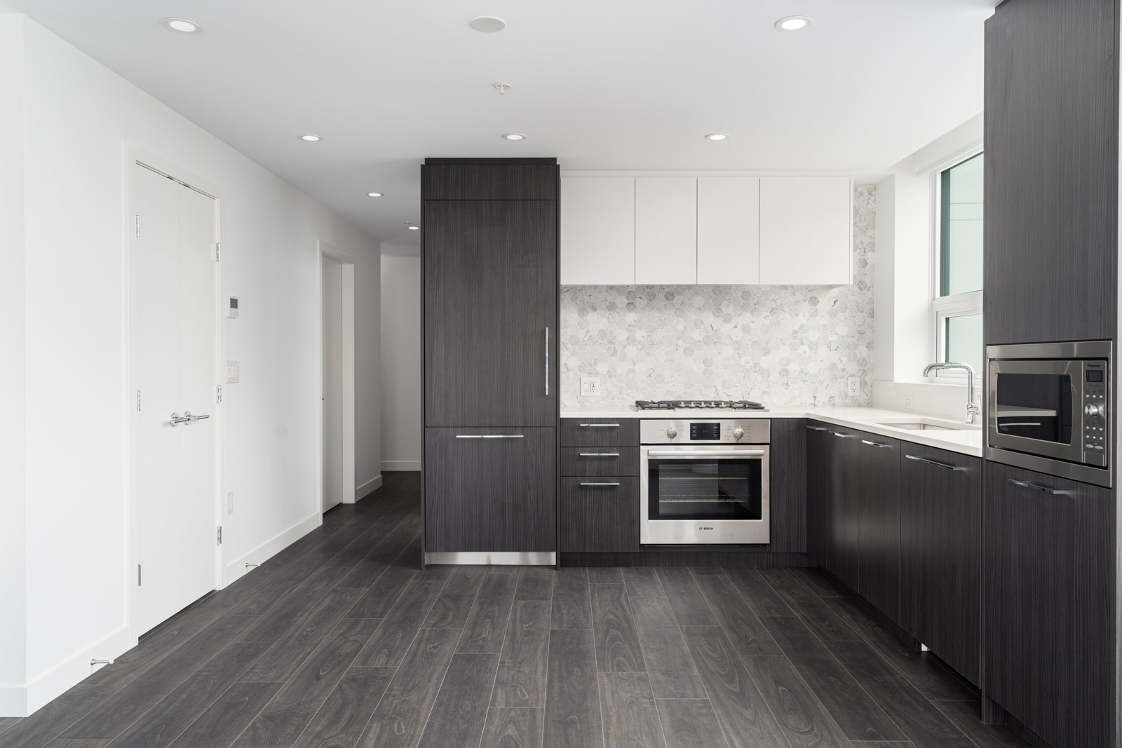 Modern kitchen with dark wood cabinets, white upper cabinets, built-in appliances, light countertops, and a patterned backsplash, featuring dark wood flooring and ample lighting.
