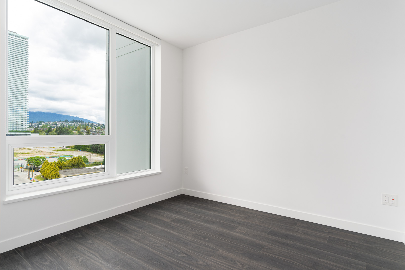 Empty room with white walls, large window, and dark wood flooring; urban and mountain view visible outside.