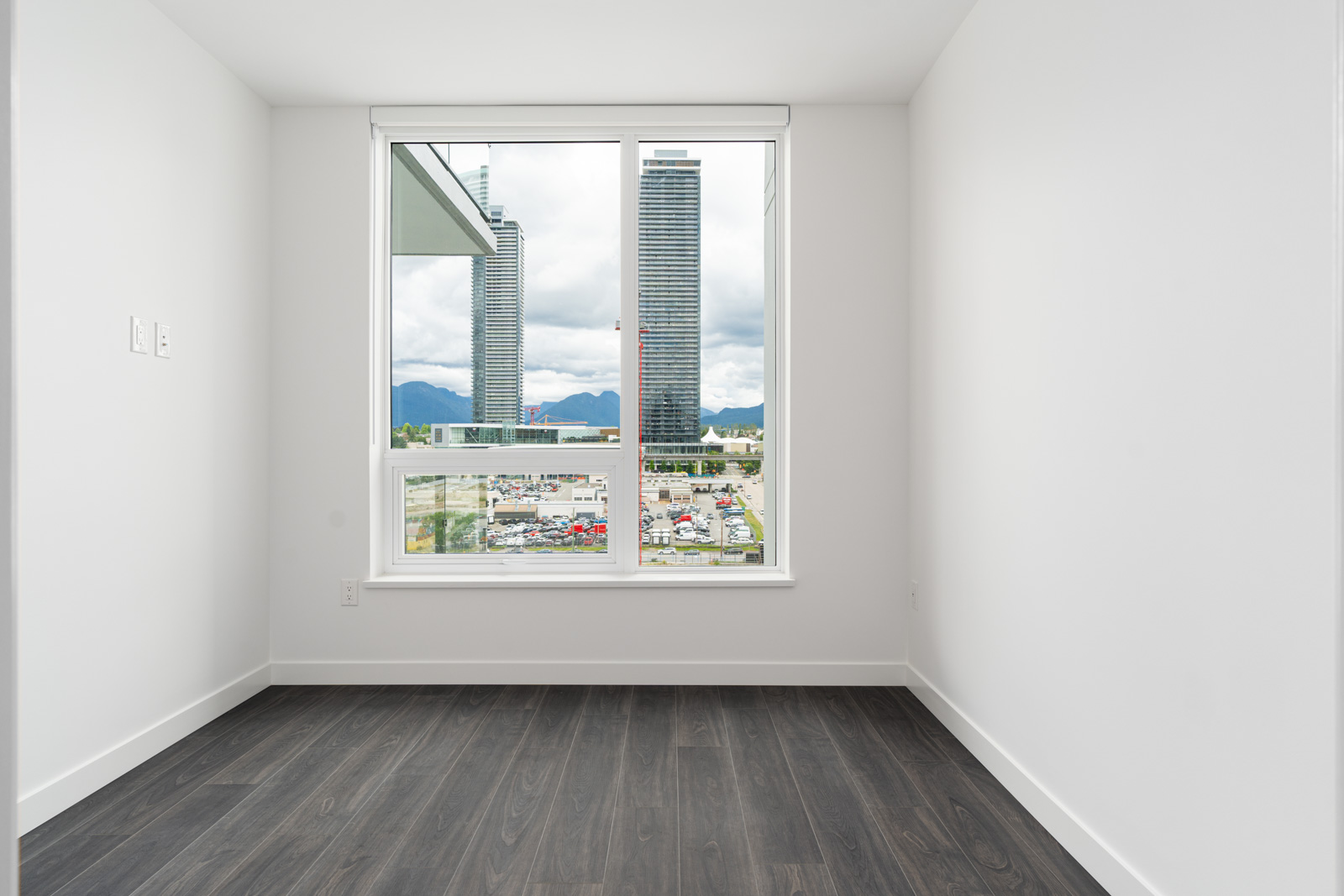 Empty white room with dark wood flooring and a large window overlooking city buildings and mountains under a cloudy sky.