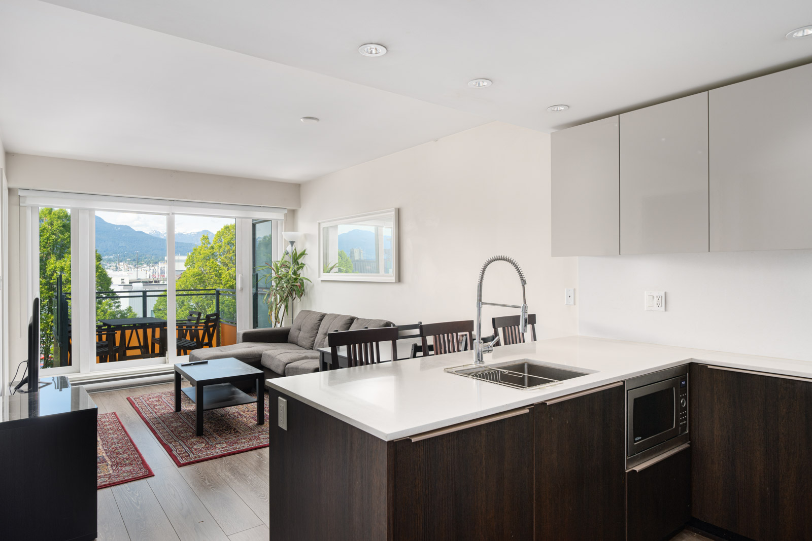 Modern open-plan apartment with a kitchen island, living area, and dining table, featuring large windows and a balcony with mountain and city views.
