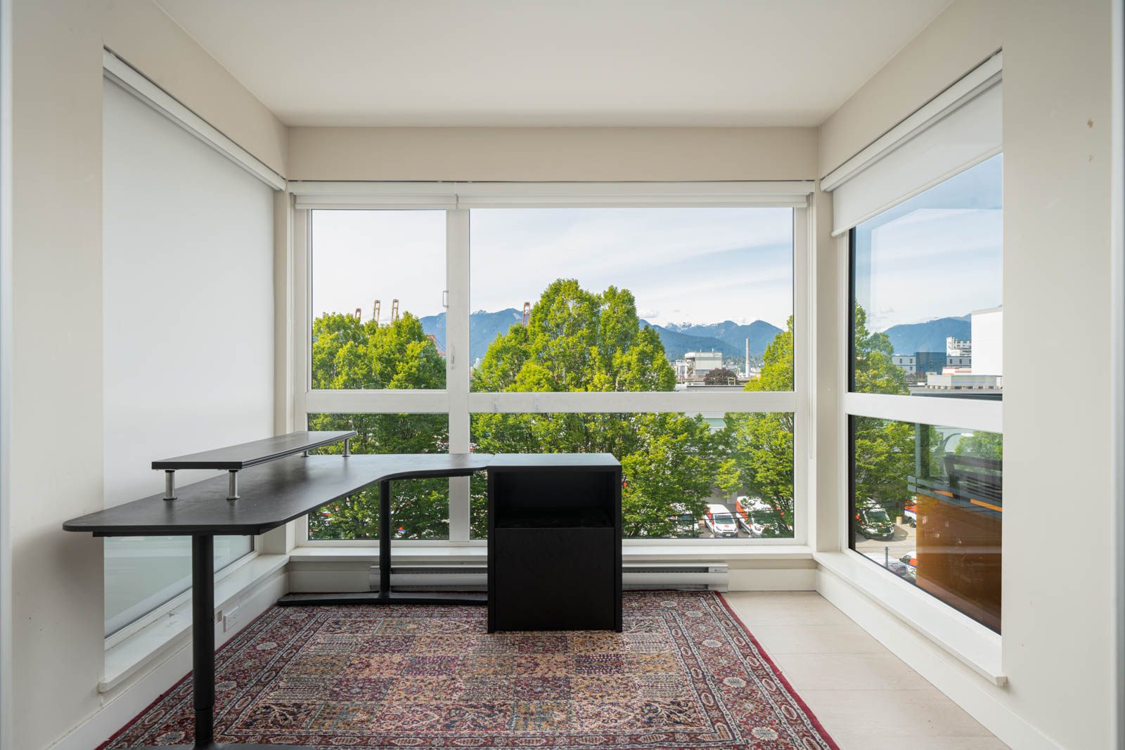 Modern corner office with large windows, black L-shaped desk, black chair, and patterned rug; trees and mountains visible outside.