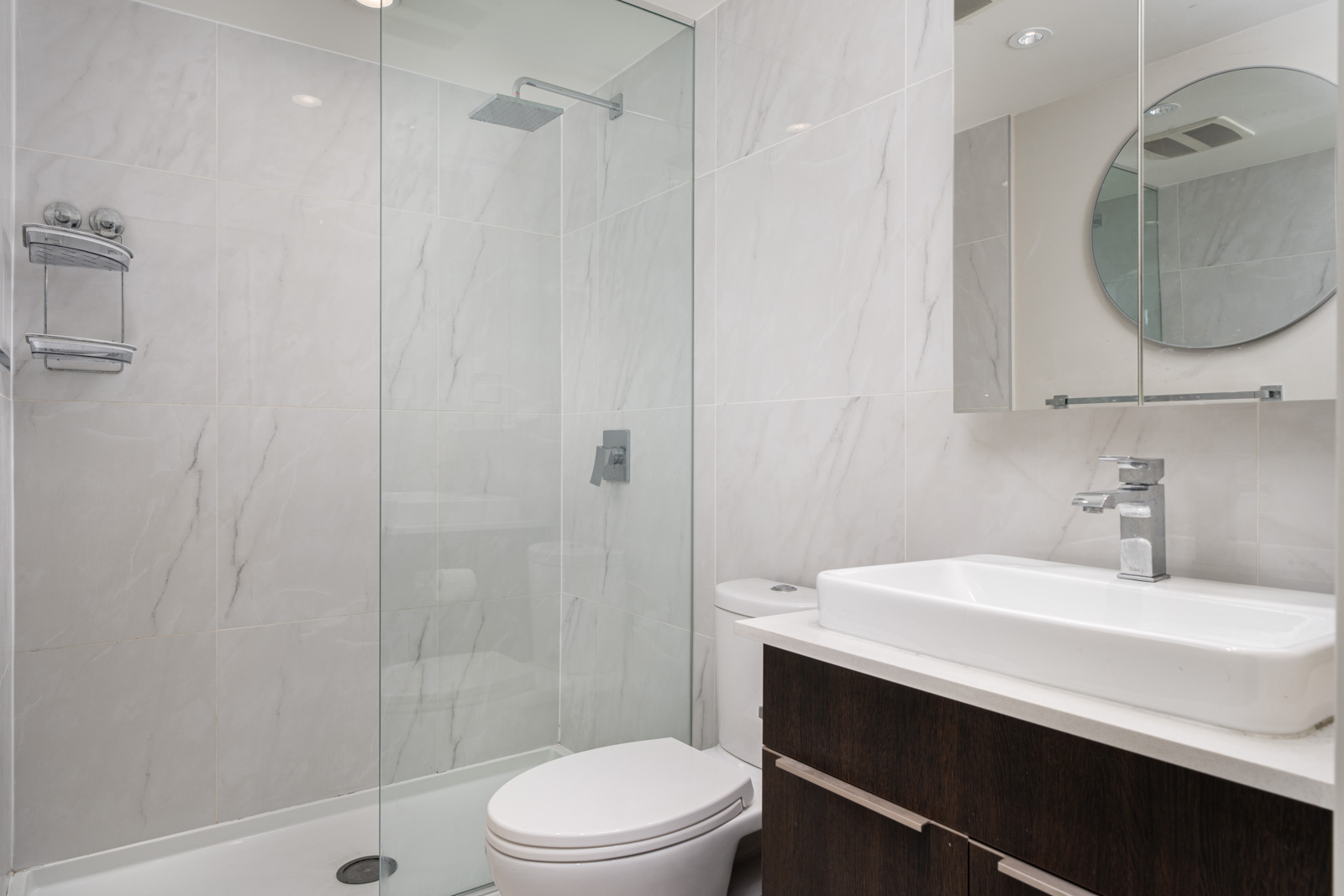 Modern bathroom with glass-enclosed shower, wall-mounted showerhead, white toilet, rectangular sink on dark vanity, and round mirror above the sink. Marble tile on the walls.