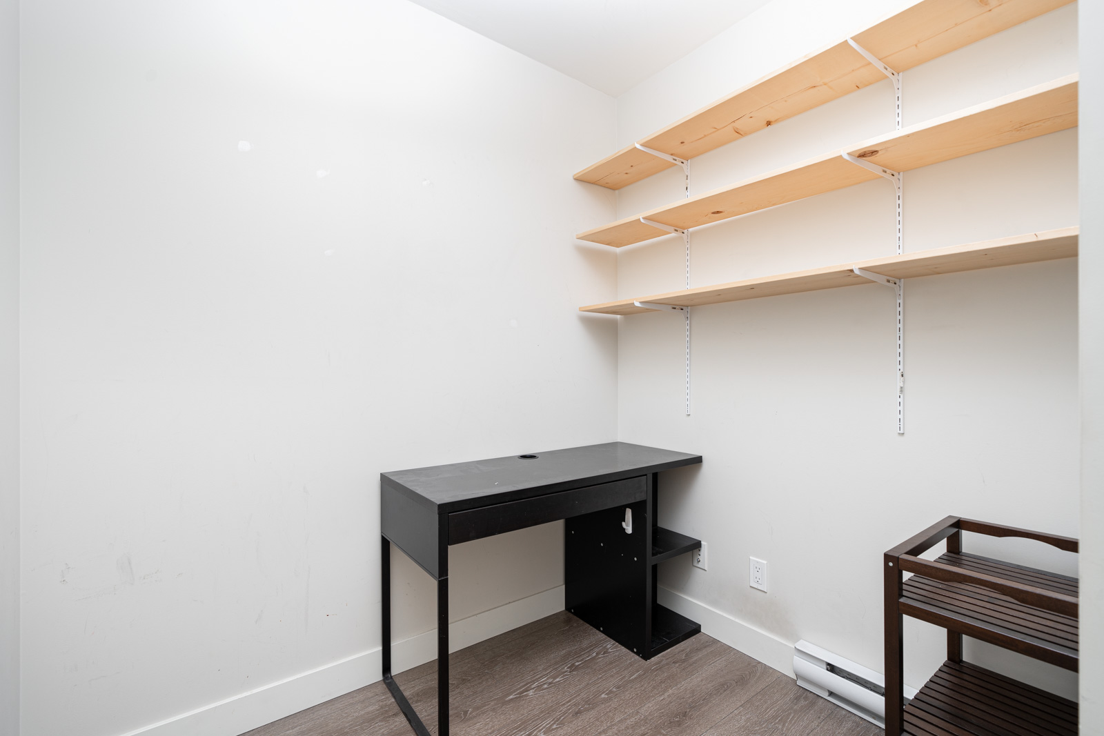 Small, empty room with a black desk, wooden shelves on the wall, and a small brown shelving unit on the right, with white walls and wood flooring.