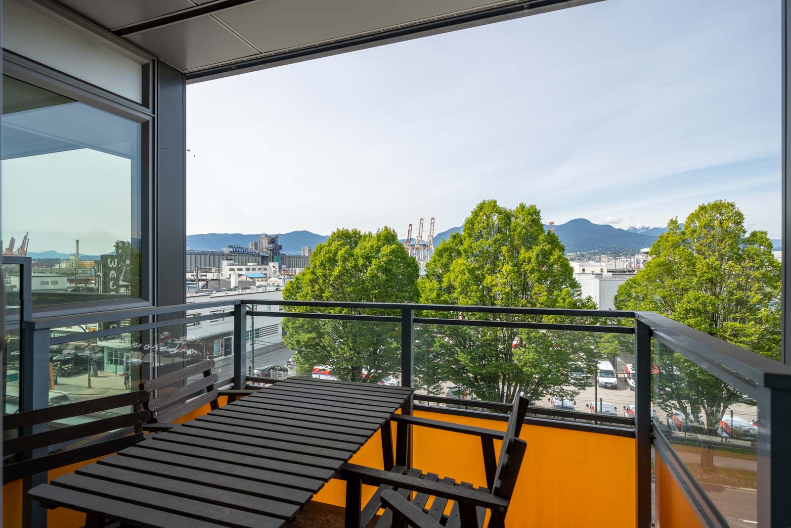 A balcony with a black outdoor table and chairs overlooks trees, a street, industrial buildings, and distant mountains under a clear sky.