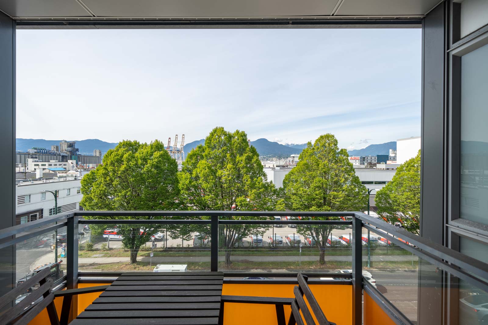 A balcony with a small table and railing overlooks green trees, a street with parked cars, industrial buildings, and distant mountains under a clear sky.
