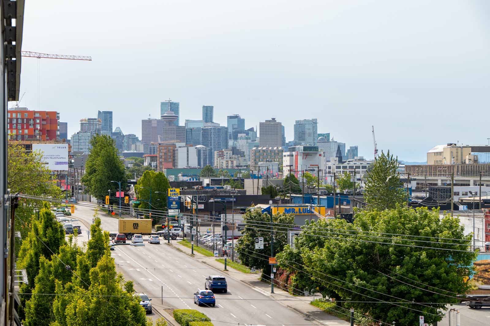 View of a busy urban street with cars and trees in the foreground and a city skyline with tall buildings in the background under a cloudy sky.