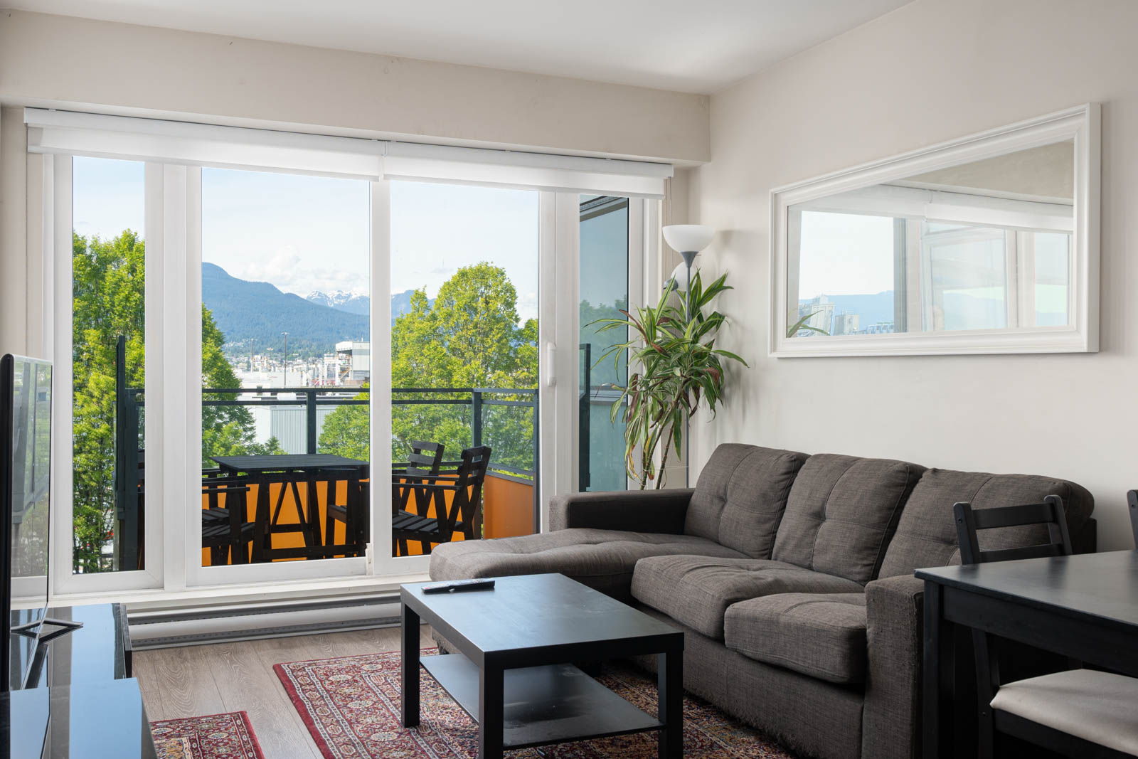 A modern living room with a gray sofa, black coffee table, and large glass doors leading to a balcony with outdoor seating and a view of mountains and trees.