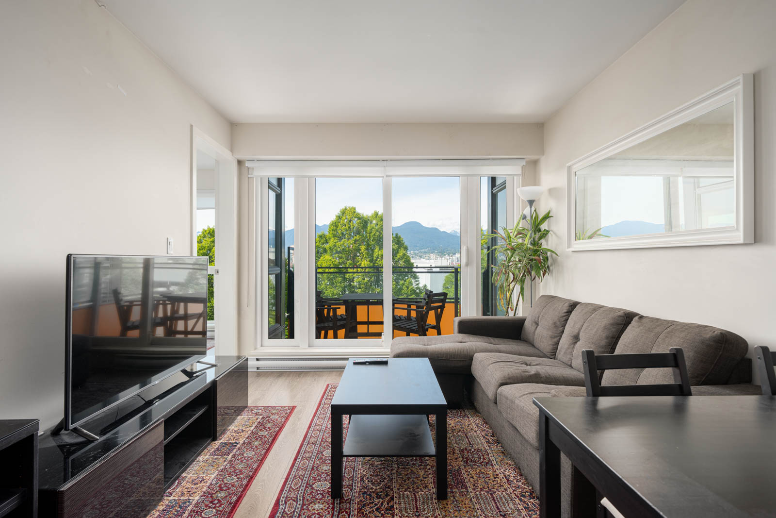 Modern living room with a gray sectional sofa, TV, coffee table, and large windows leading to a balcony with outdoor seating and a mountain view.
