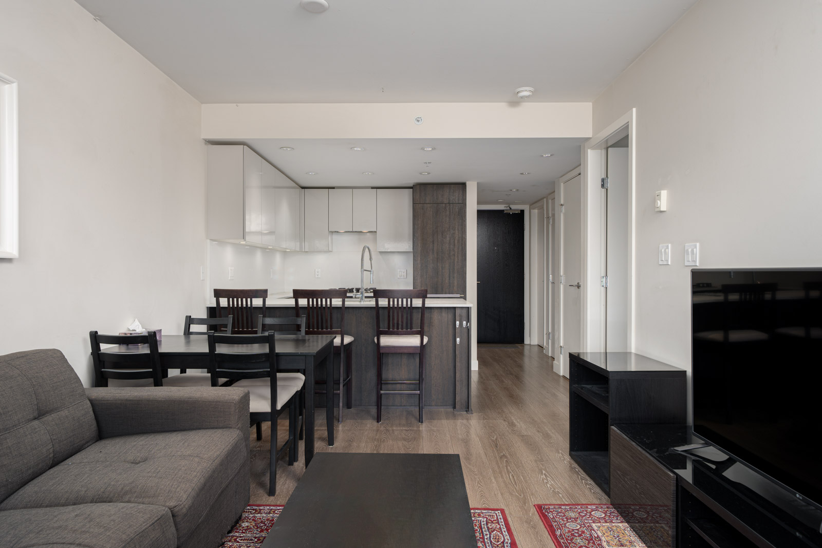 Modern apartment interior with a gray sofa, dining table with chairs, open kitchen, wood flooring, and a TV stand against a white wall.