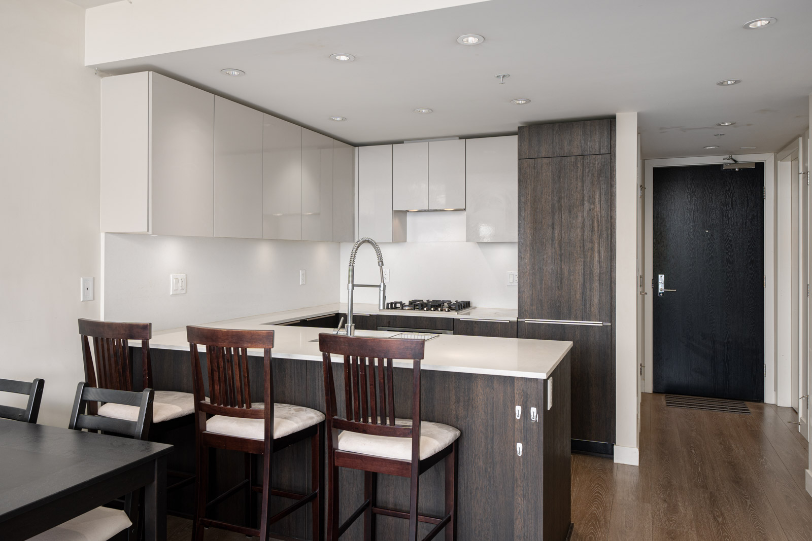 Modern kitchen with dark wood cabinets, white countertops, and three bar stools at the island; a black door and dining table are visible in the background.