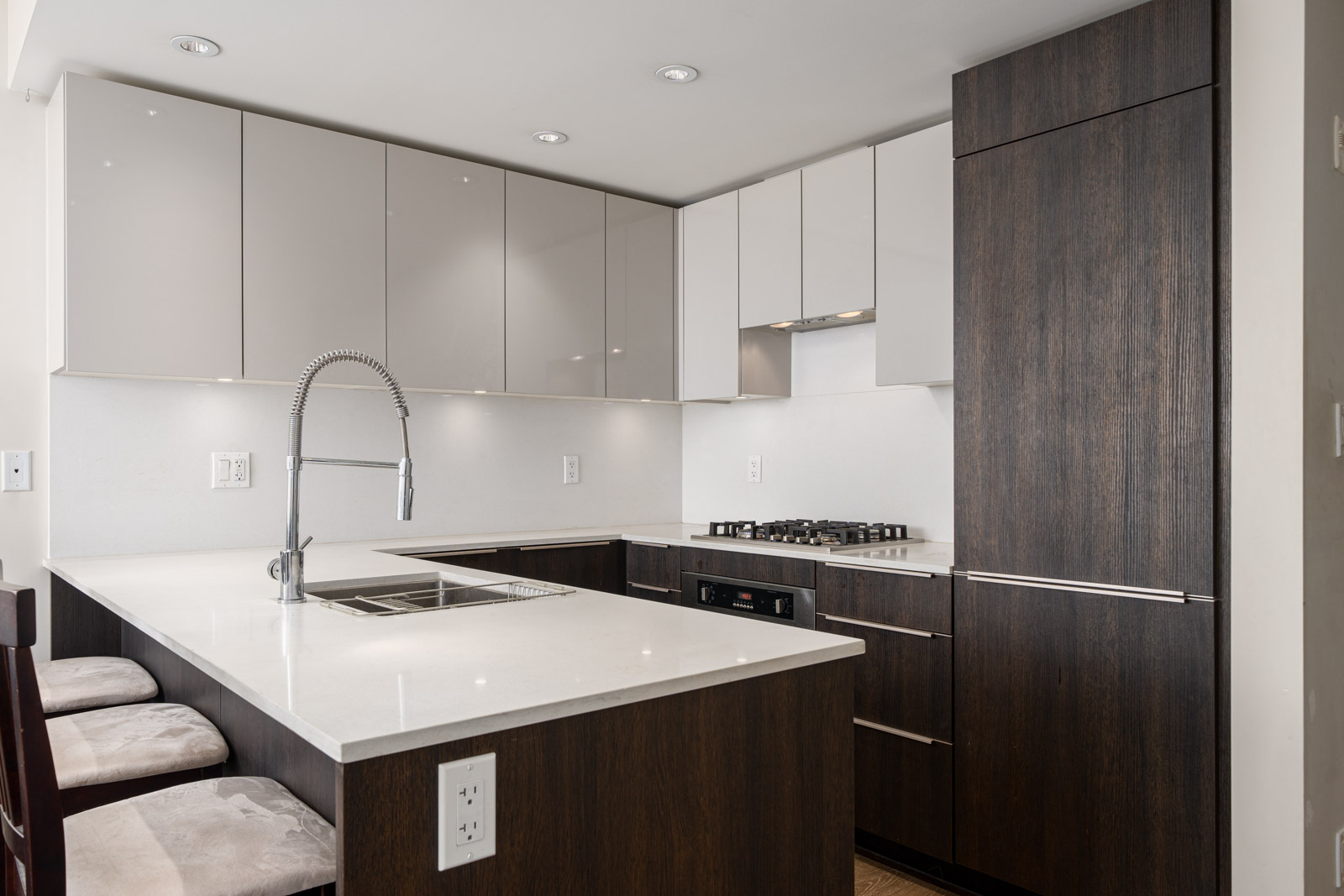 Modern kitchen with dark wood cabinetry, white countertops, an integrated sink with a tall faucet, gas cooktop, and built-in appliances. Three cushioned chairs are placed at the counter.
