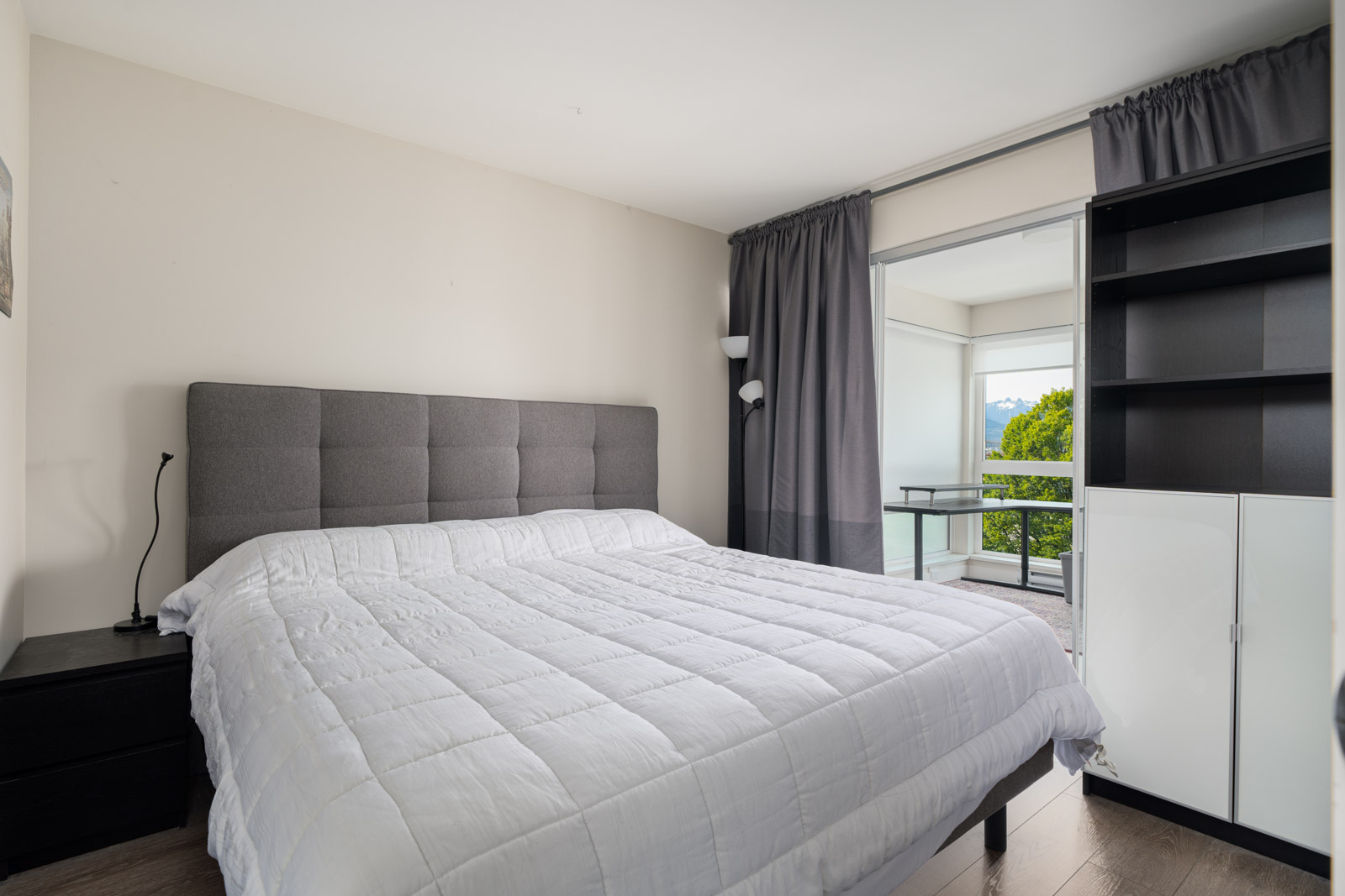 Modern bedroom with a large bed, gray headboard, dark nightstands, floor lamp, shelving unit, and sliding door opening to a balcony with greenery visible outside.