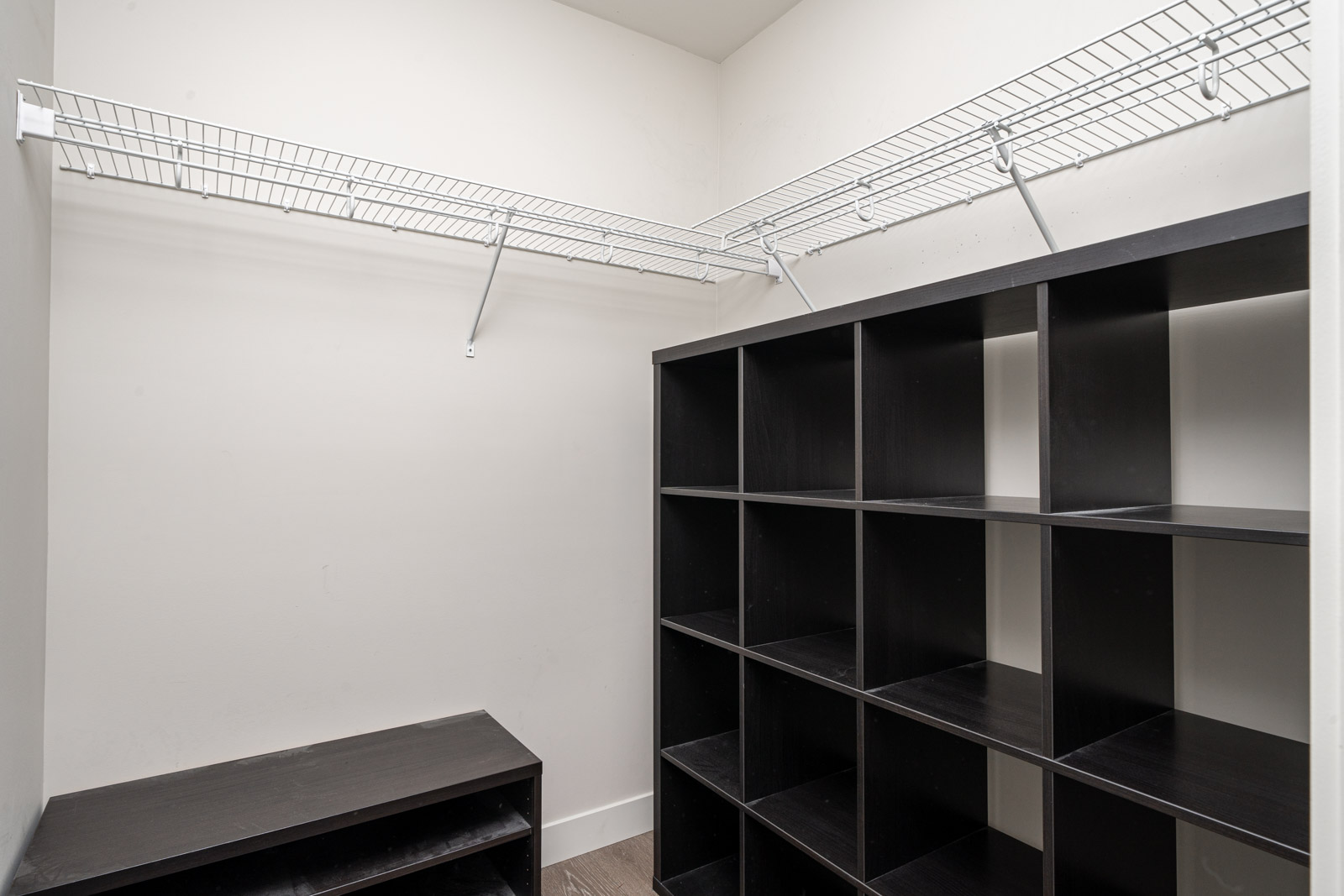 Empty walk-in closet with white wire shelves on the walls and a black cubby storage unit and bench on the floor.
