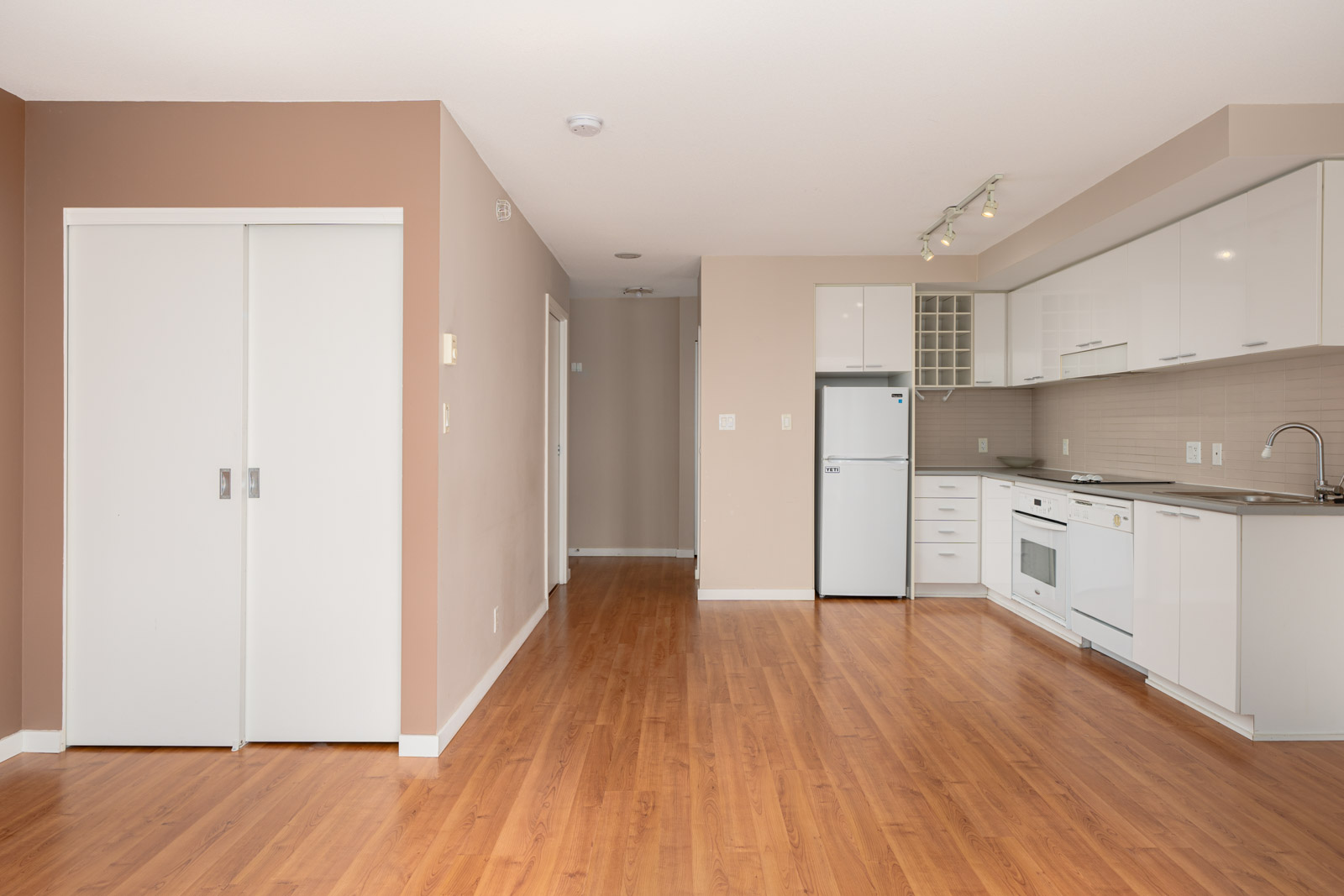 Empty modern kitchen with white cabinets, white appliances, light brown walls, wooden flooring, and a hallway leading to the back. Double white sliding doors are on the left.