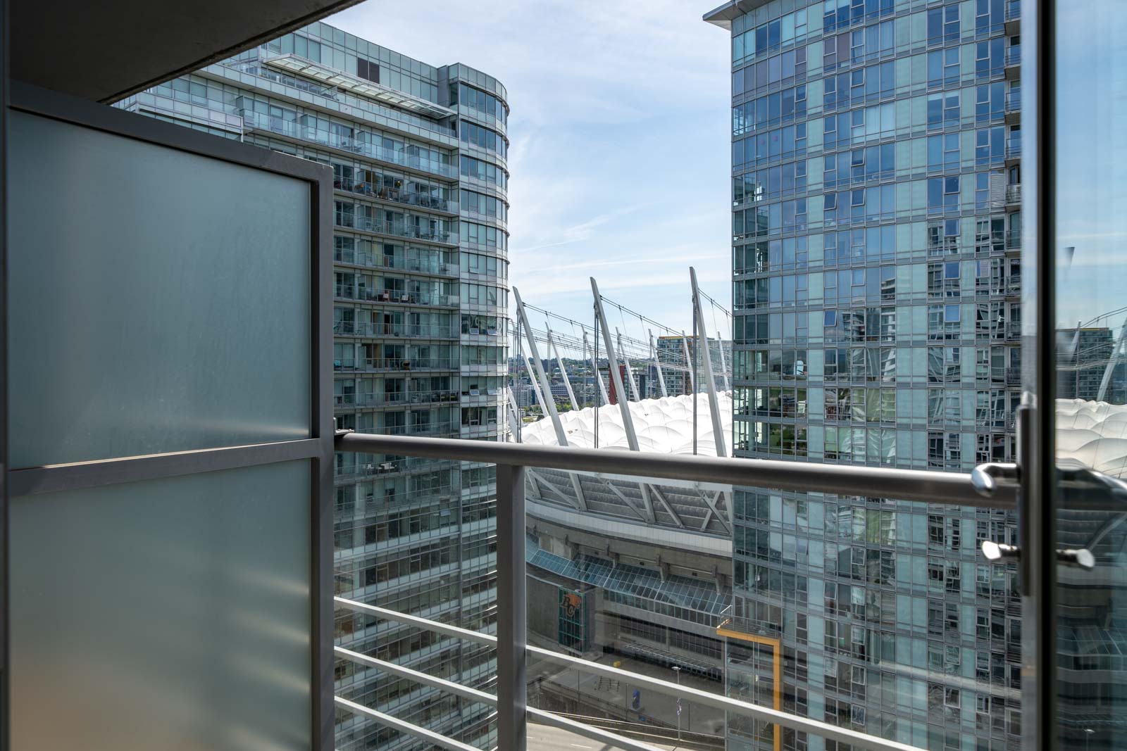 View from a balcony with glass railing, showing modern high-rise buildings and a stadium with white roof structure in the background under a clear sky.