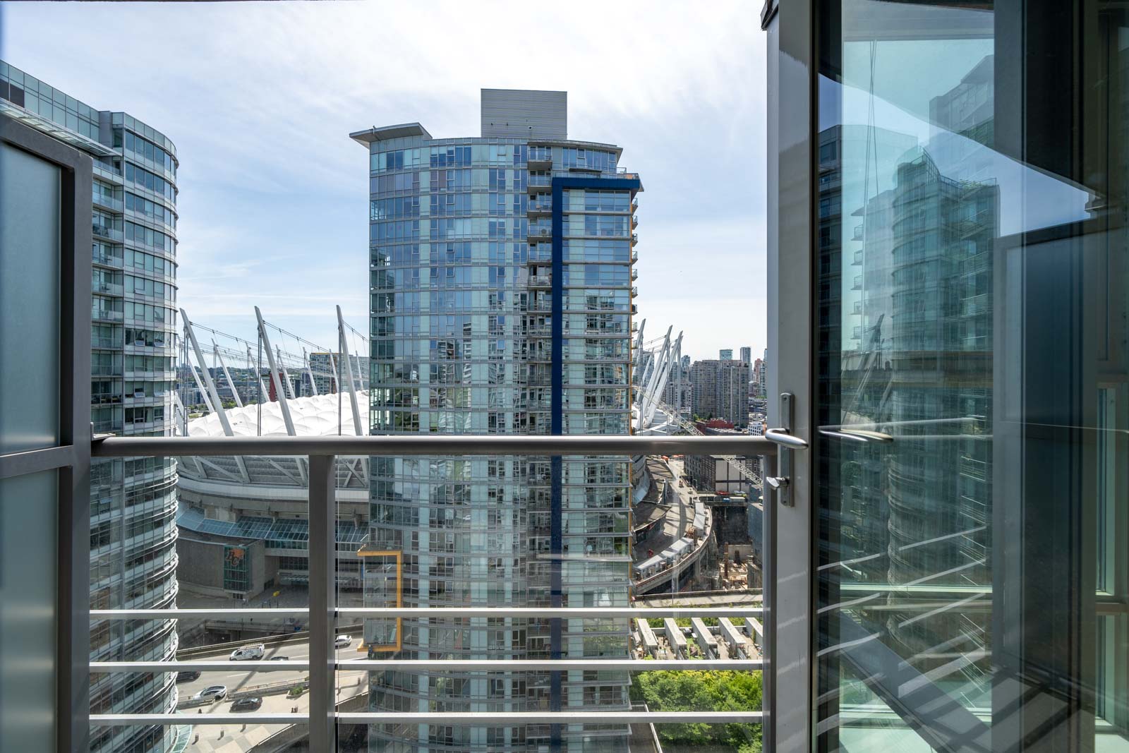 View from a balcony showing modern glass high-rise buildings, a stadium with white roof supports, city streets, and a partly cloudy sky.