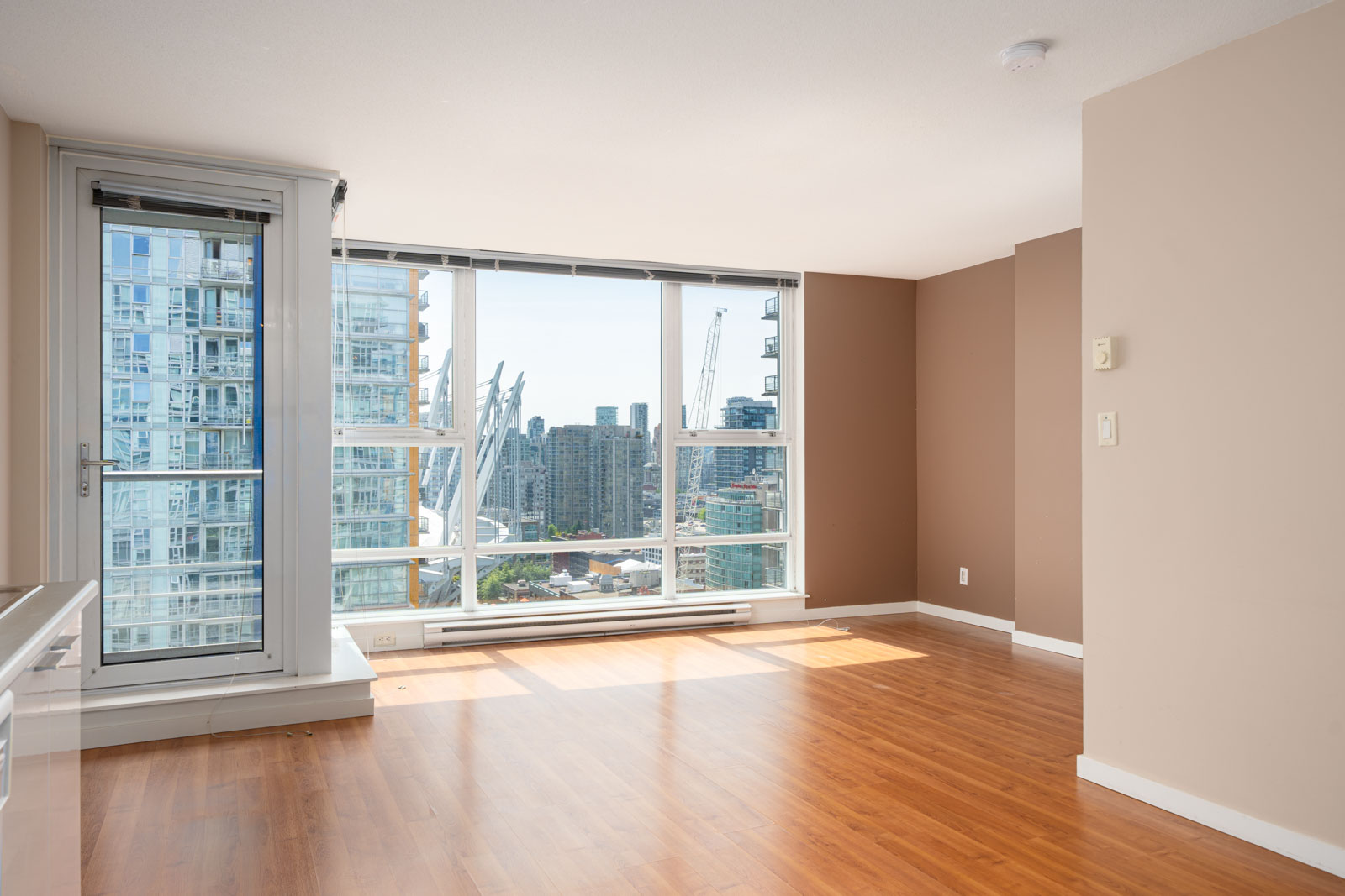 Unfurnished apartment with large windows, wood flooring, and a view of city buildings and construction cranes outside.