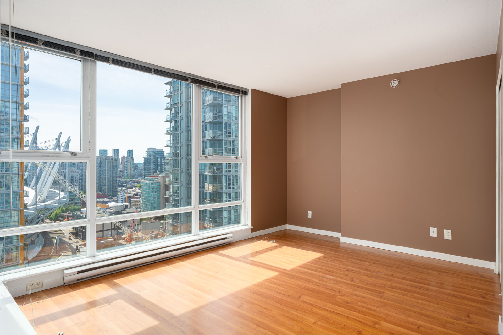 Empty room with brown walls, large windows, and hardwood floors, featuring a city skyline view with modern high-rise buildings in the background.