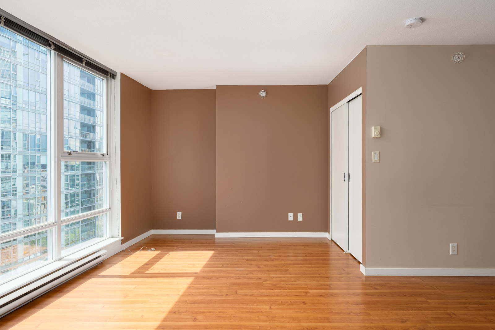 Empty room with brown walls, hardwood floor, large windows on the left, and white sliding closet doors on the right.