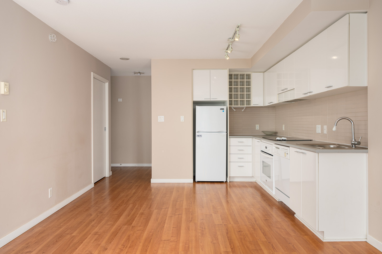 Modern kitchen with white cabinets, stainless steel sink, refrigerator, oven, and wood flooring, adjacent to an empty beige wall and hallway with a closed white door.