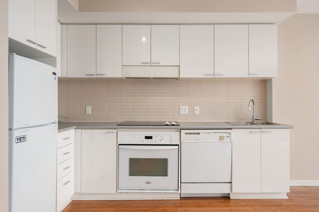 A modern kitchen with white cabinets, a refrigerator, stove, oven, dishwasher, sink, and tiled backsplash against a light beige wall.
