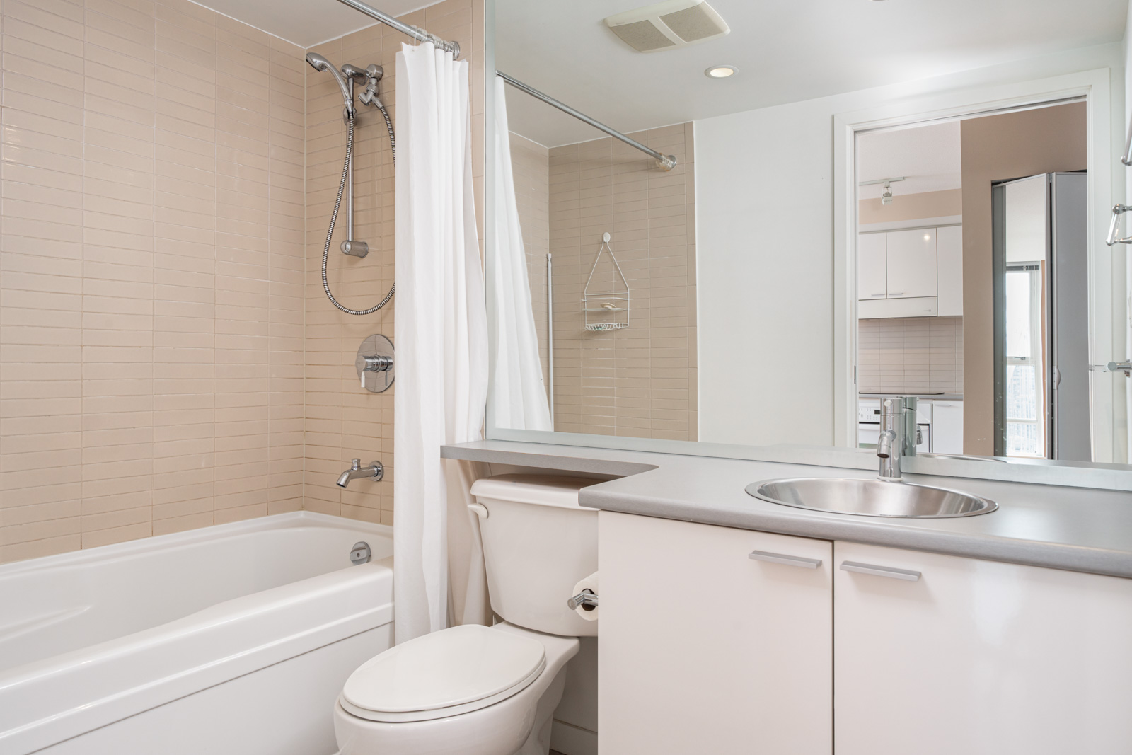 Modern bathroom with a bathtub and shower, white curtain, toilet, gray countertop with sink, large mirror, and neutral tiled wall. Doorway shows part of a kitchen.