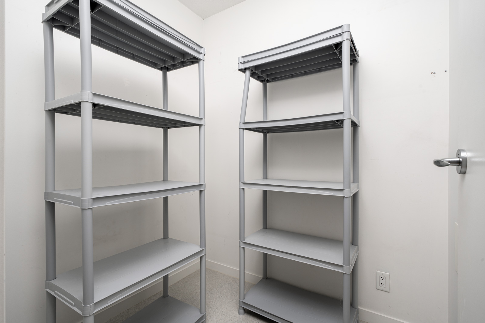 Two gray plastic shelving units stand against the walls of a small, white, empty room with a closed door and a power outlet visible near the floor.