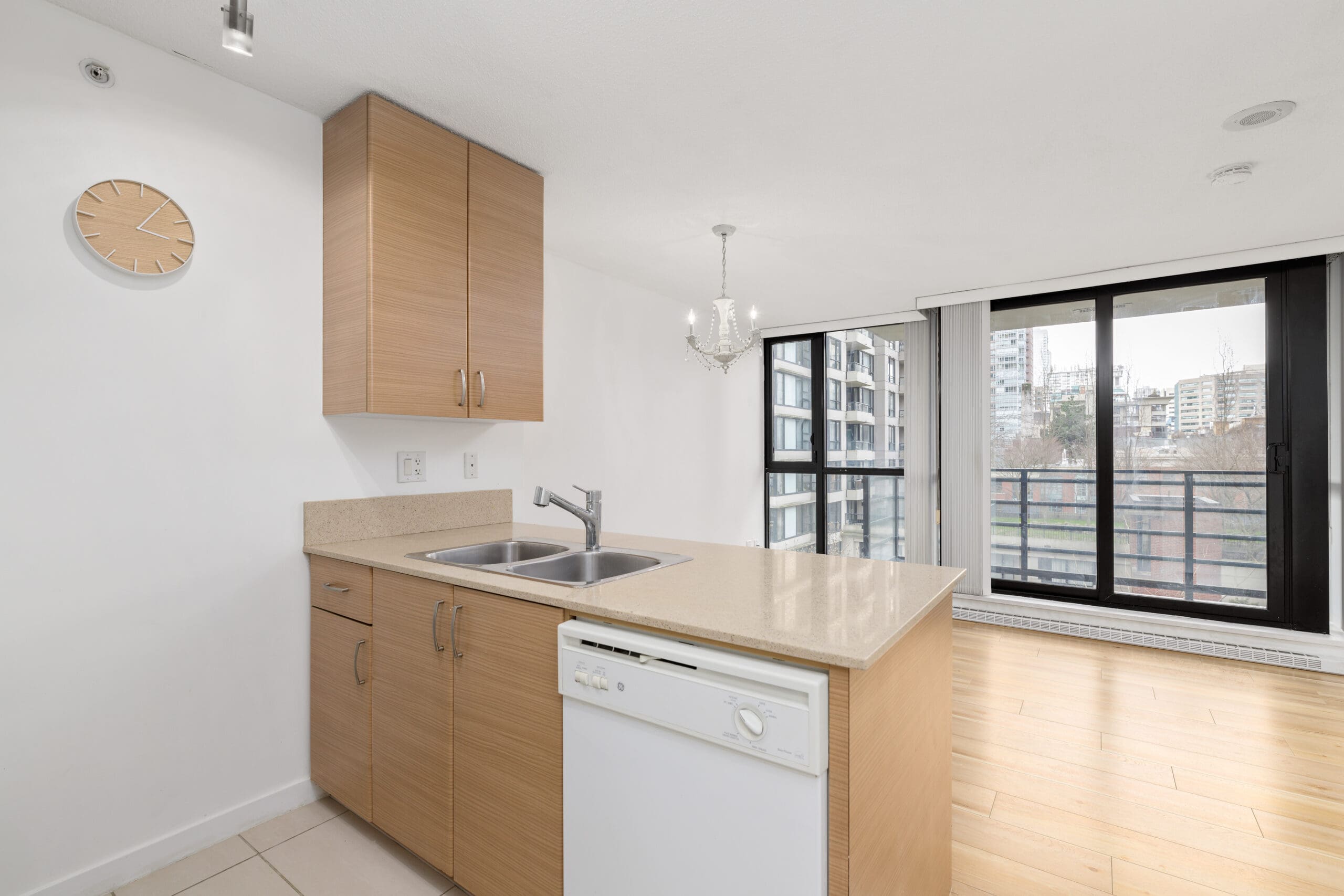 Modern kitchen with light wood cabinets, a double sink, dishwasher, and island; large windows show a city view with buildings and a balcony.