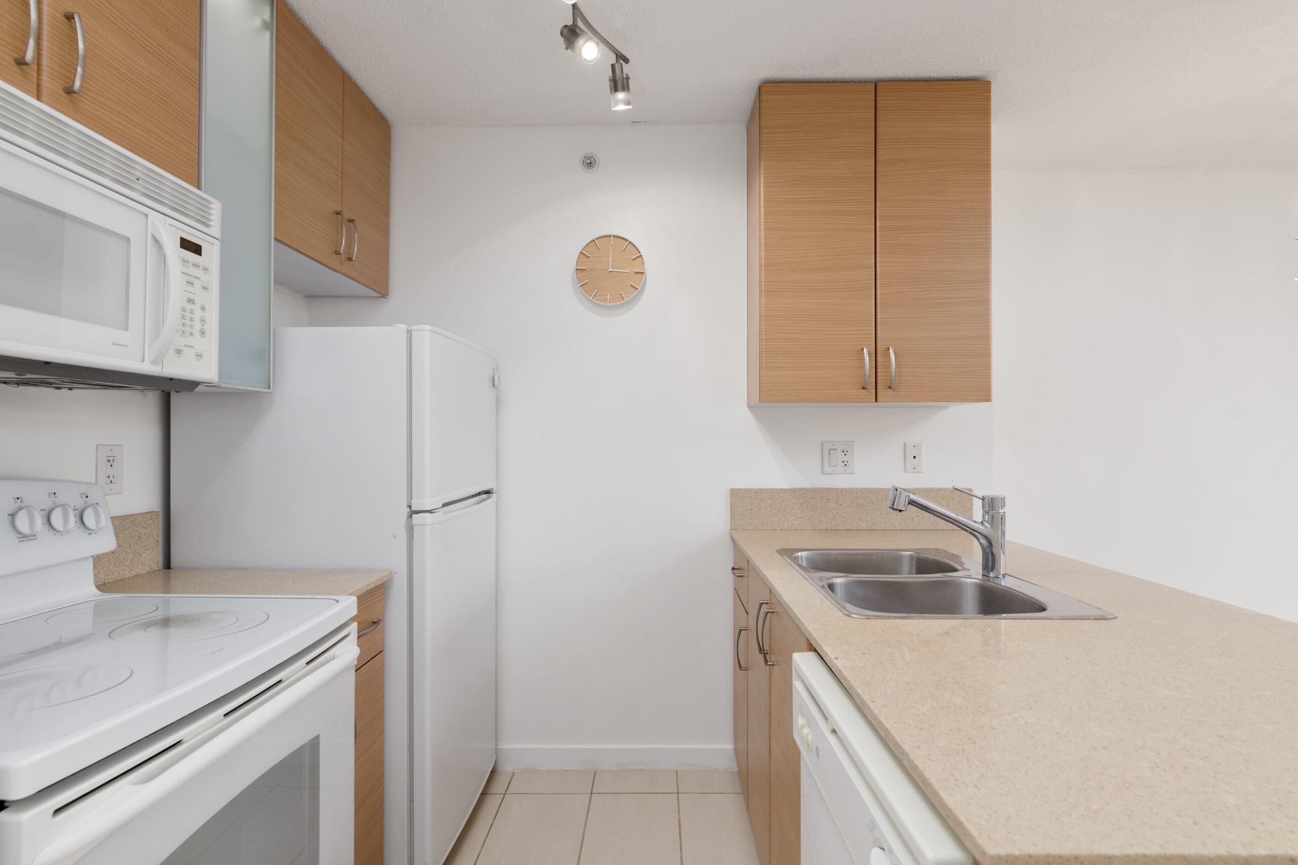 Modern kitchen with light wood cabinets, white appliances including stove, microwave, and refrigerator, double sink, and a wall clock above the counter.