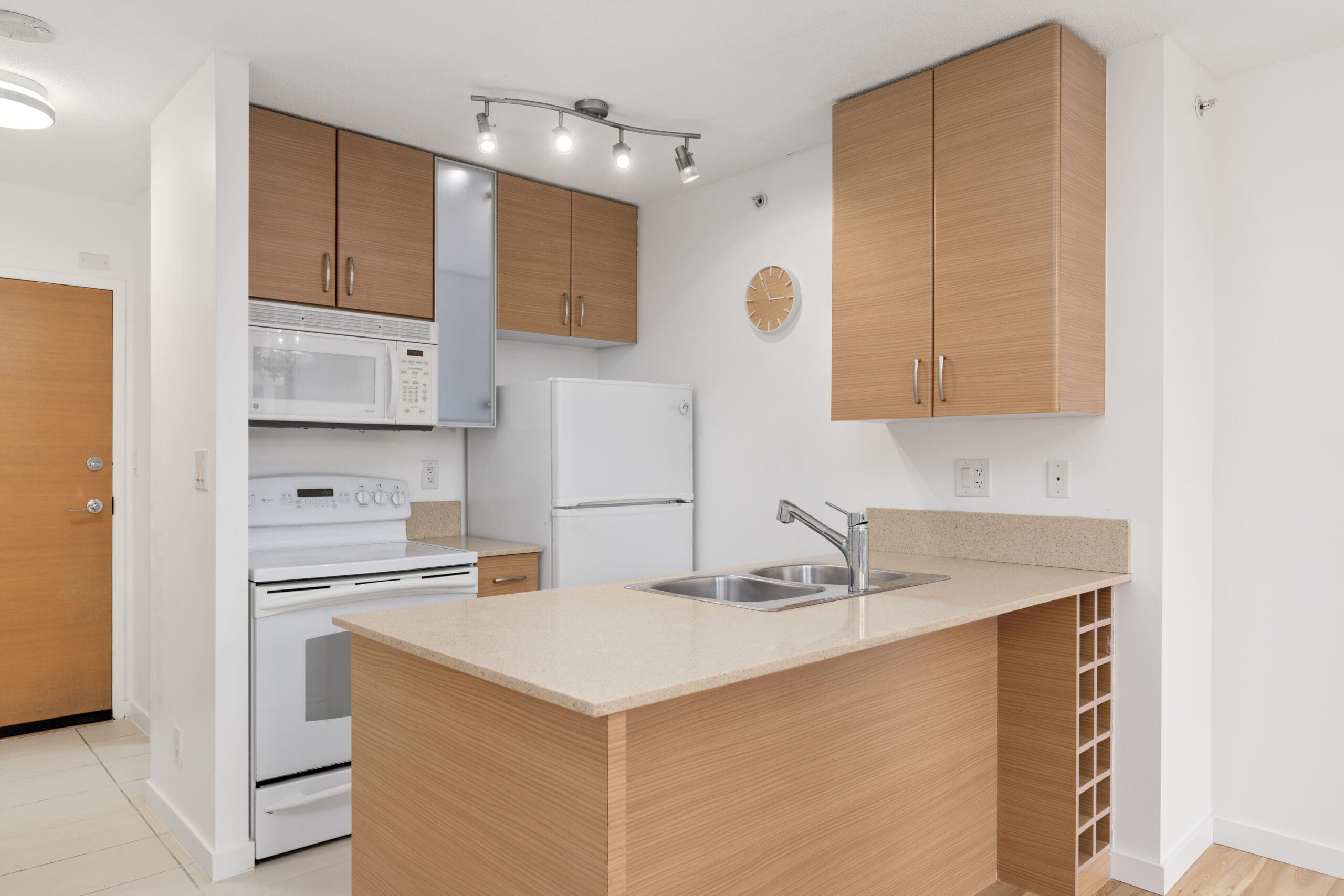 Modern kitchen with light wood cabinets, white appliances, a double sink, overhead lighting, and a small wine rack built into the island.
