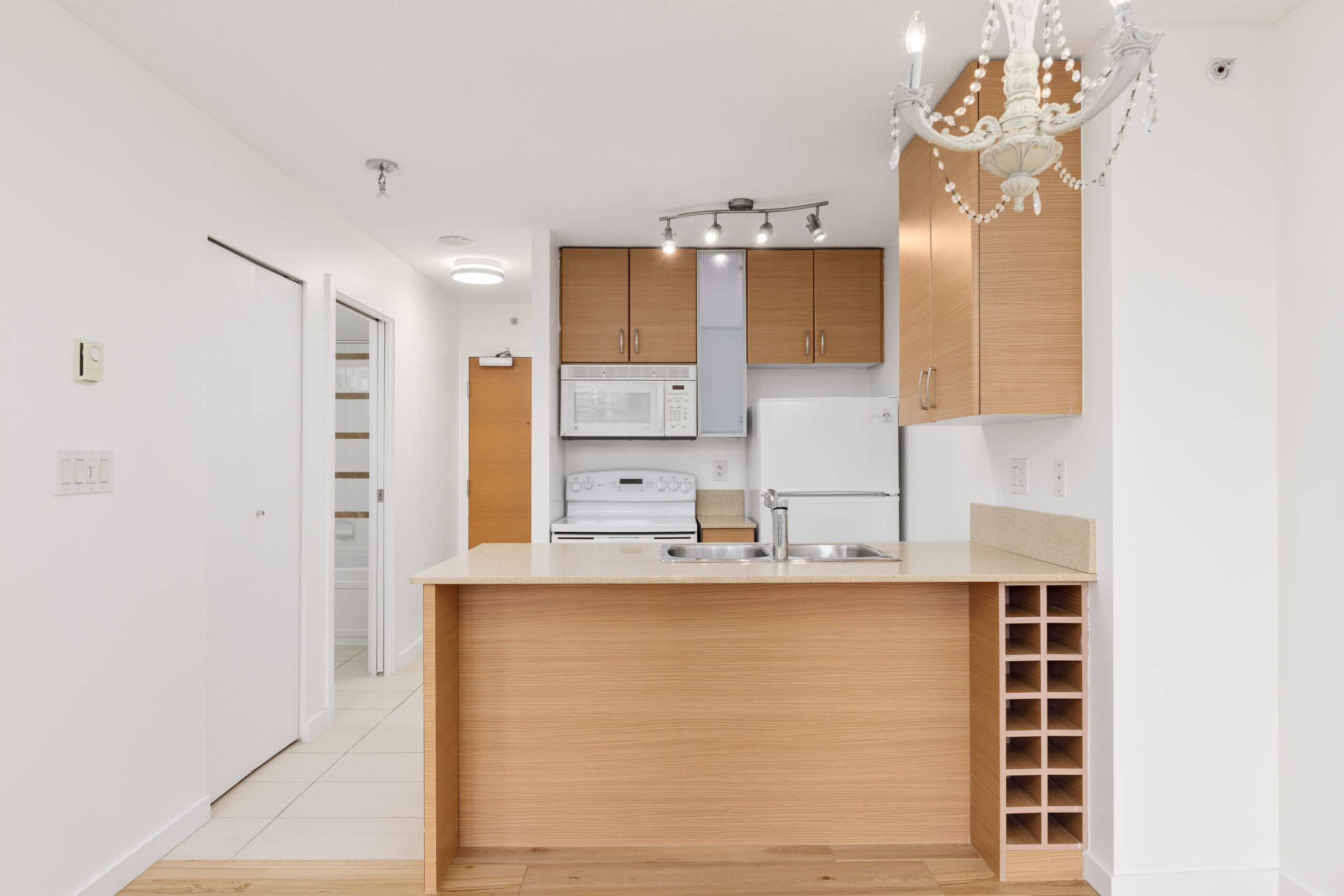 Modern kitchen with light wood cabinets, white appliances, an island with a sink, built-in wine rack, and a chandelier.