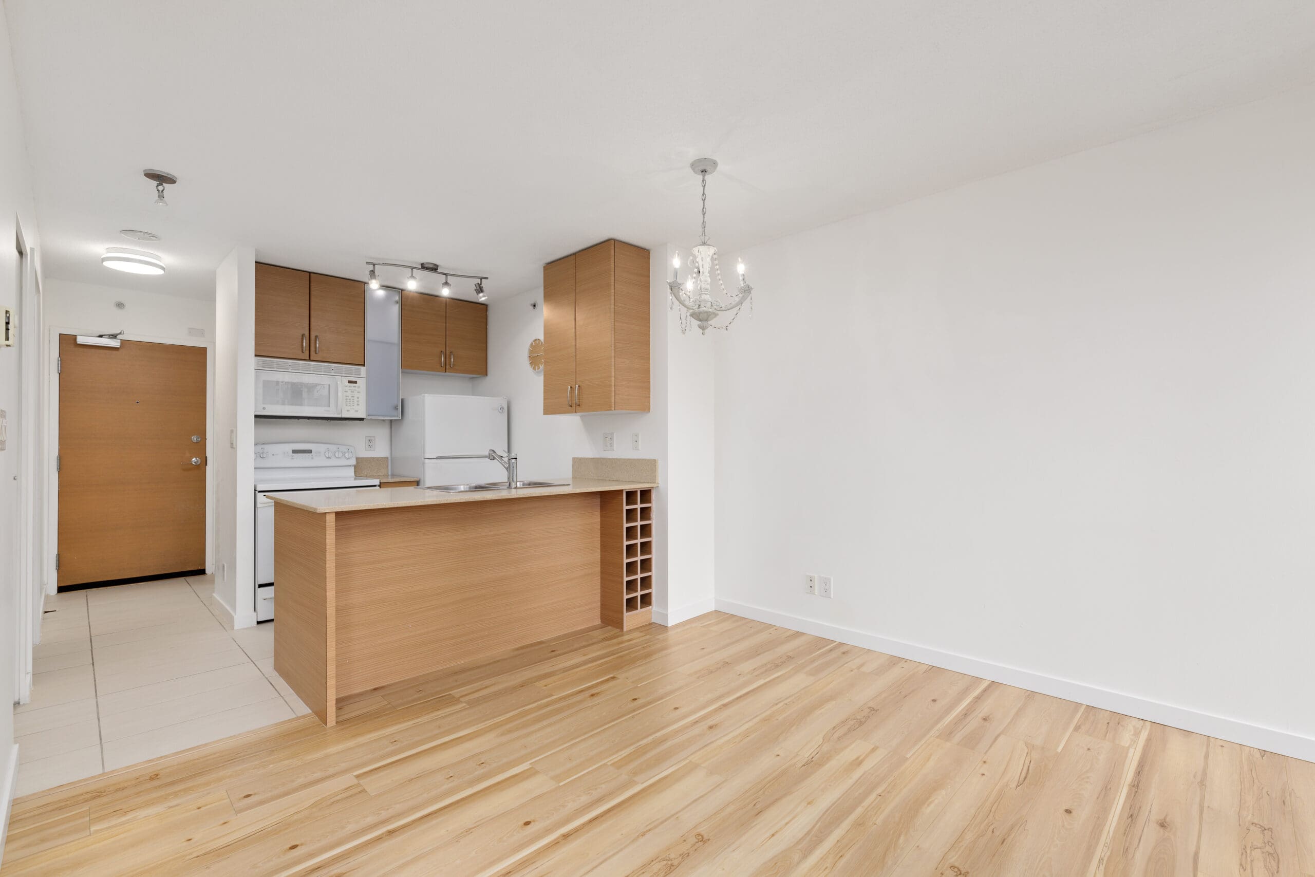 Modern kitchen with wooden cabinets, white appliances, and an island adjoining an open living area with wood floors and a chandelier.