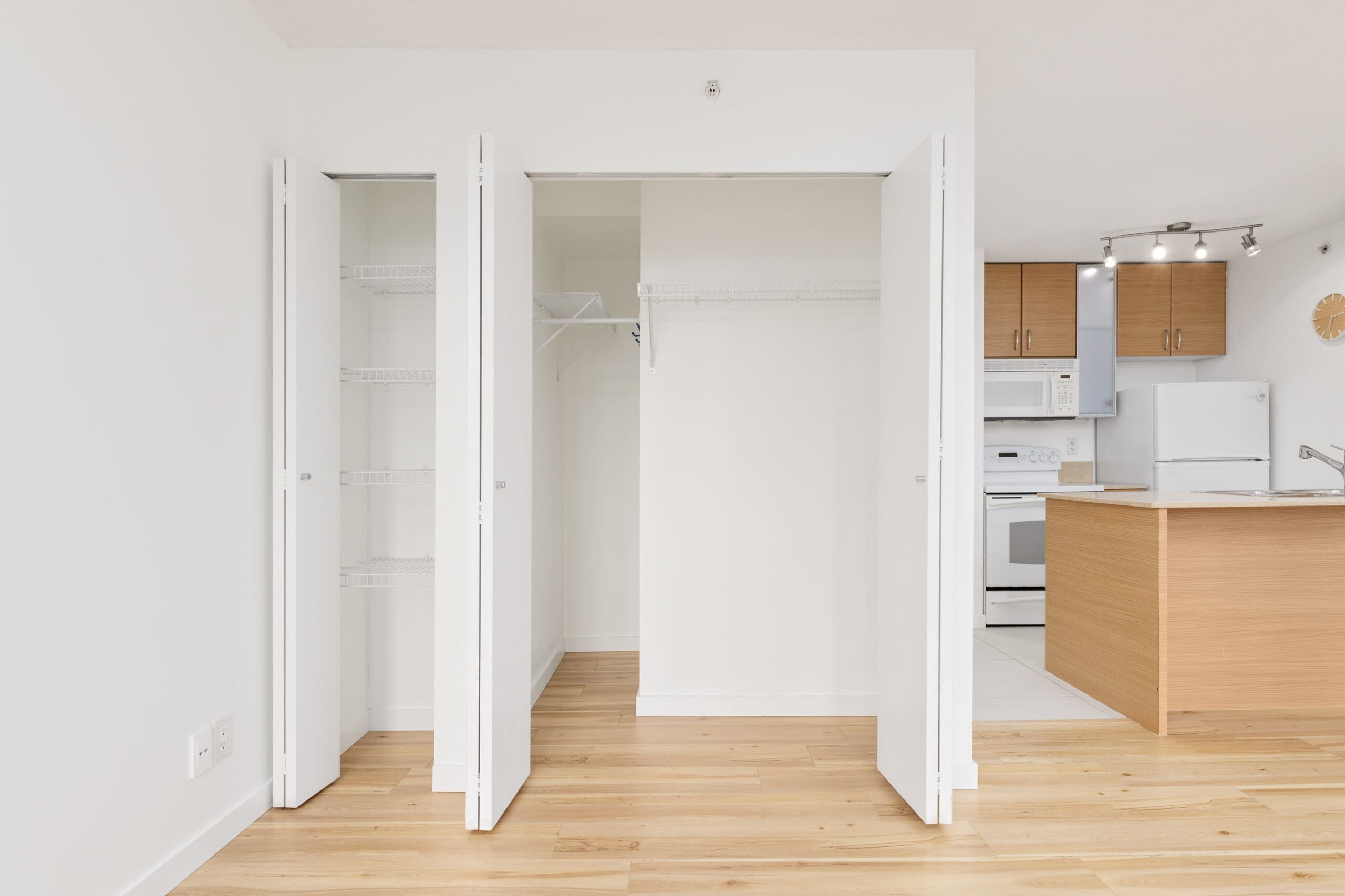 A modern apartment interior with open double closets, light wood flooring, white walls, and a view of a kitchen with wooden cabinets and white appliances.