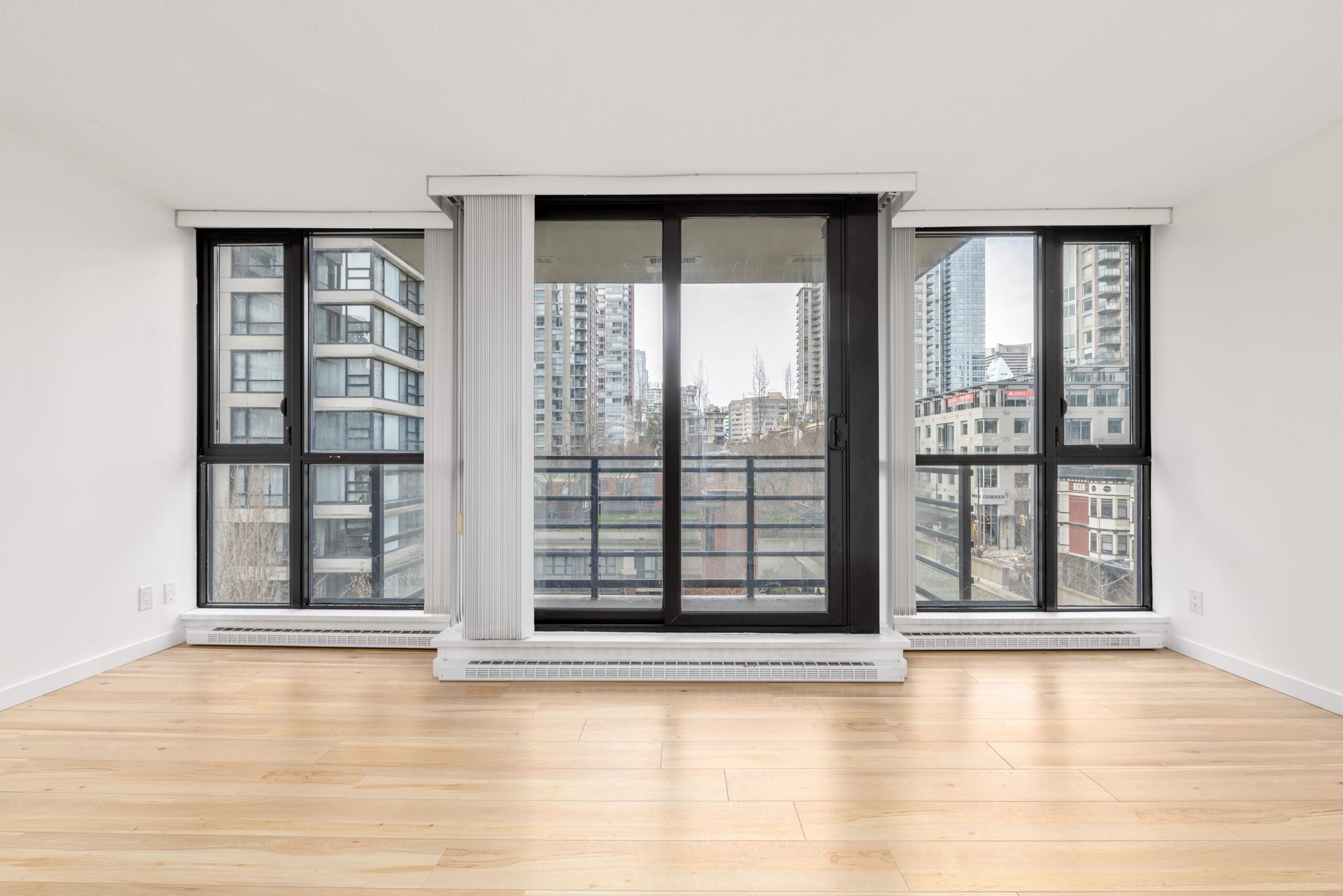 Empty modern room with light wood floors, large windows, and sliding glass doors overlooking tall city buildings.
