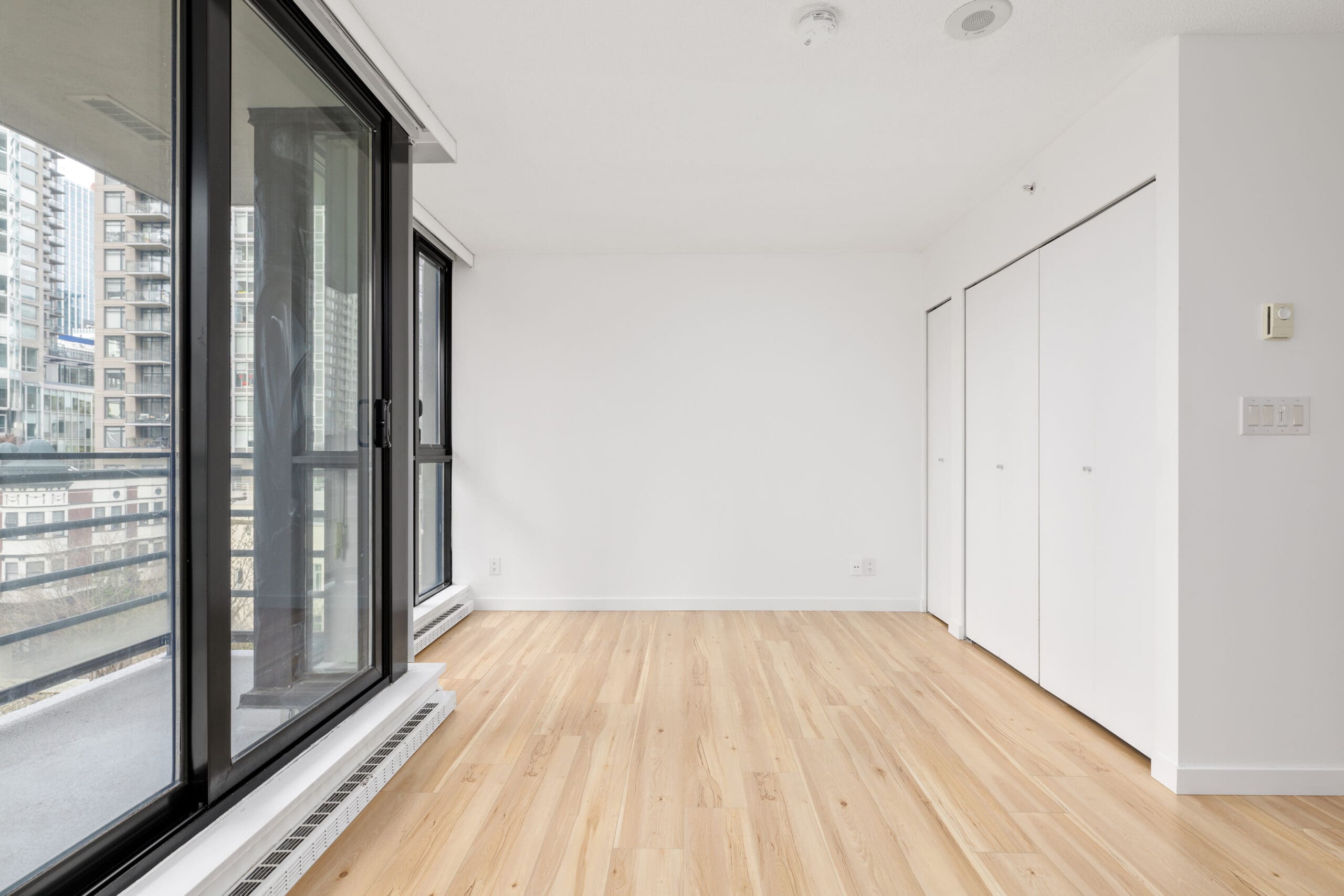 Unfurnished room with light wood flooring, large floor-to-ceiling windows, white walls, and double closet doors in a modern high-rise building.