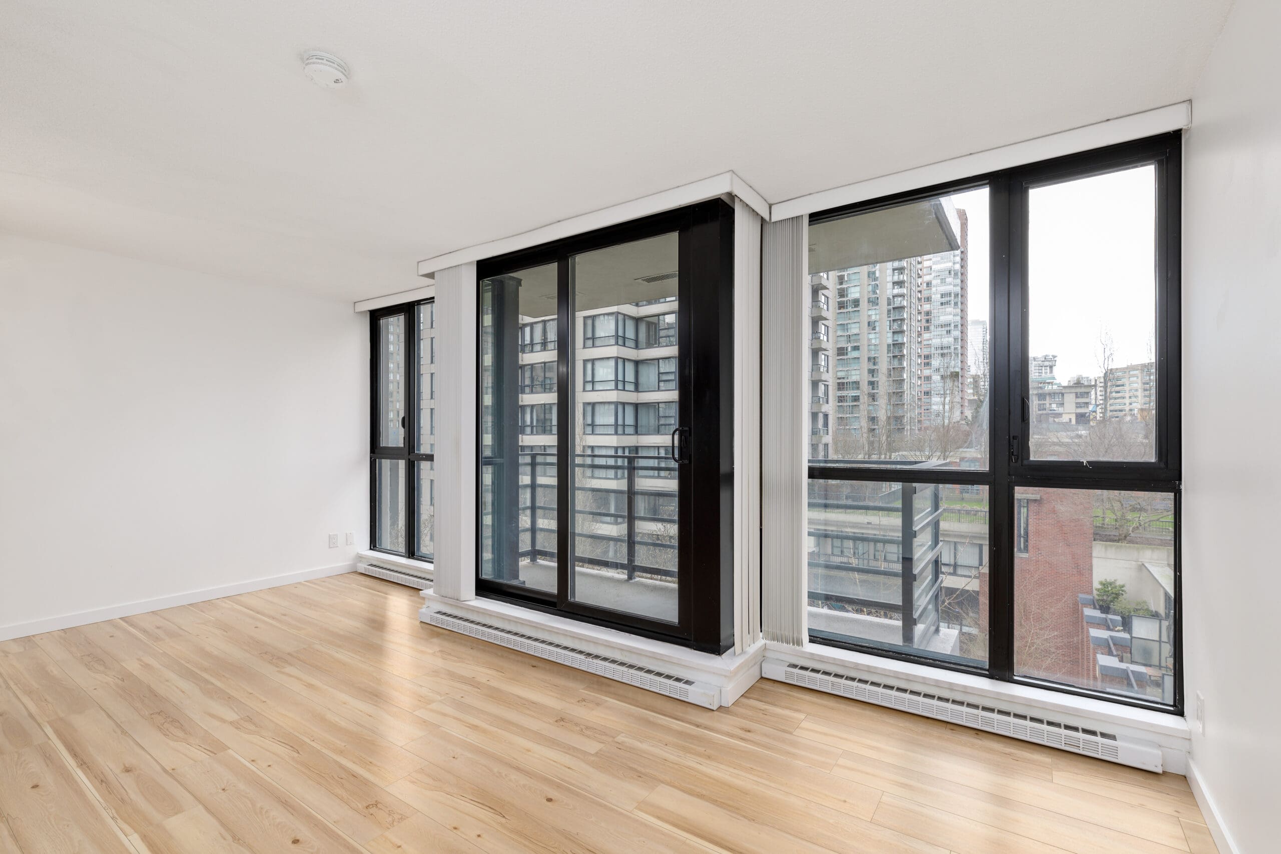 Unfurnished room with light wood flooring, large windows, and a glass door leading to a balcony overlooking modern high-rise buildings.