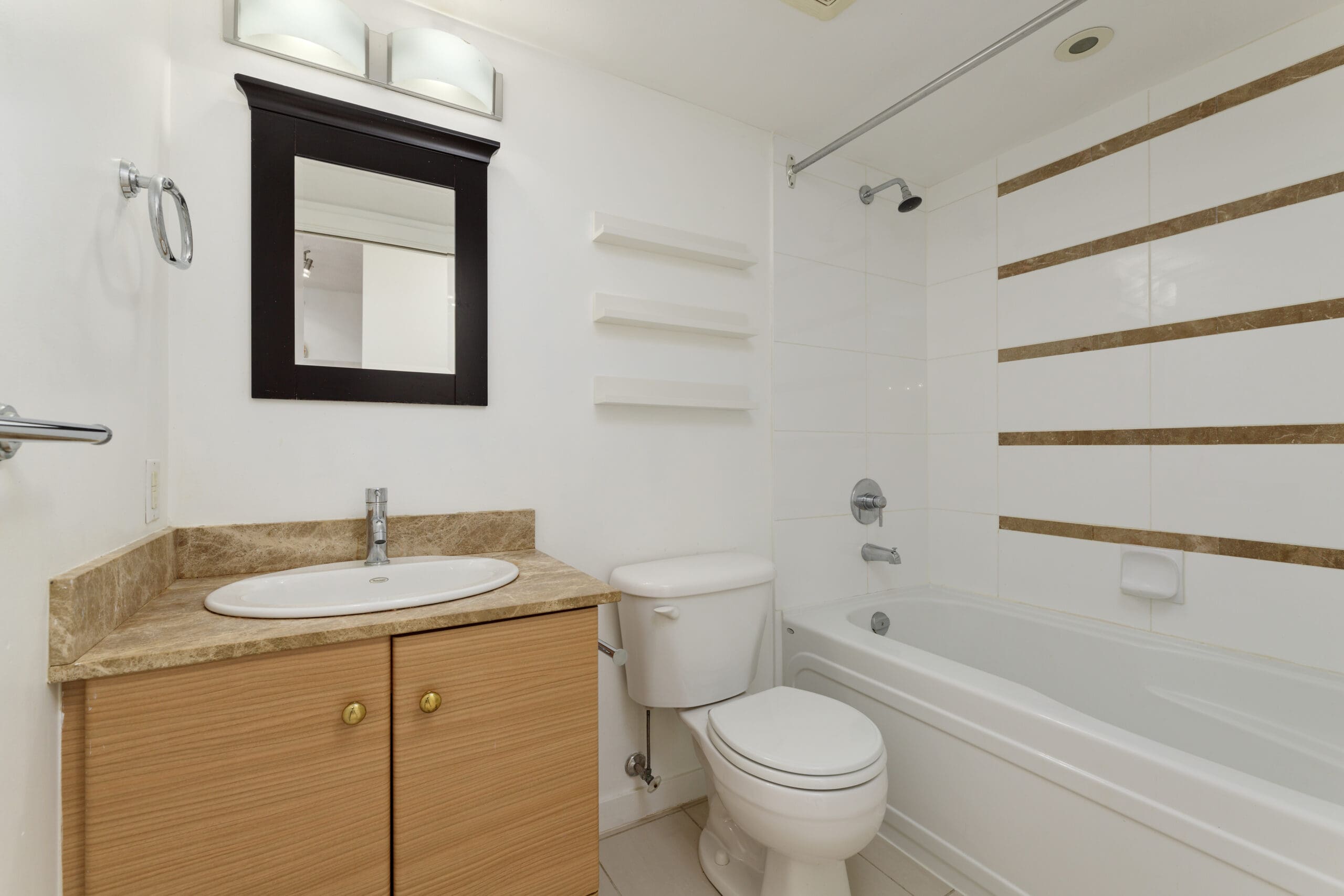 Modern bathroom with a wooden vanity, sink, mirror, toilet, and a bathtub with white tiles and brown accent stripes. Three wall shelves are mounted above the toilet.