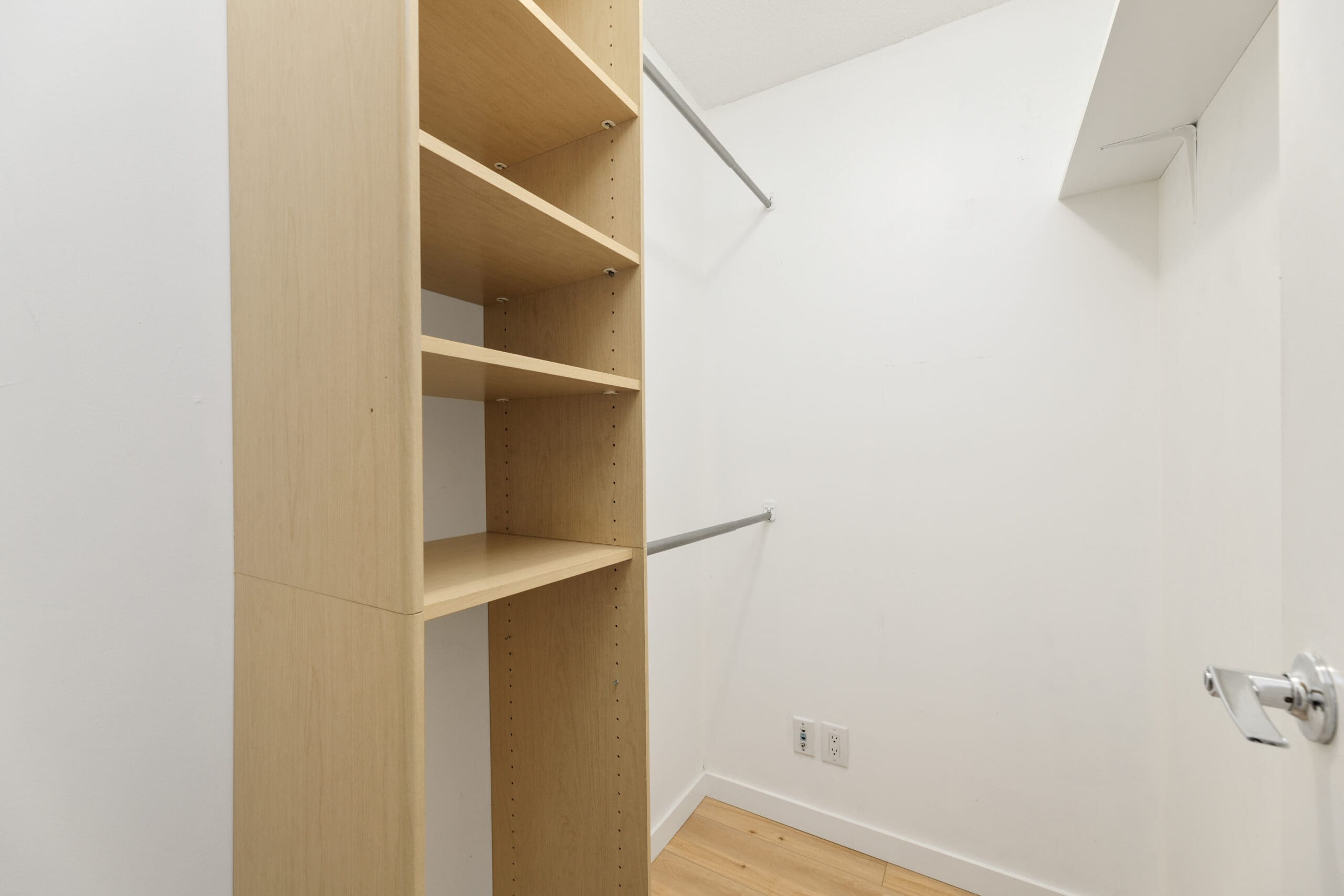 Empty walk-in closet with light wood shelving unit, two metal hanging rods, white walls, and light wood flooring.