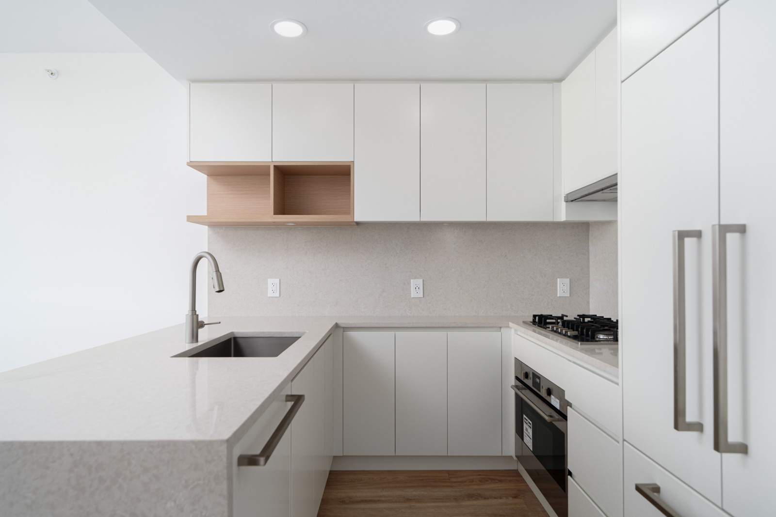 Modern kitchen with white cabinets, built-in oven, gas stovetop, stainless steel sink, light wood shelving, and quartz countertops.