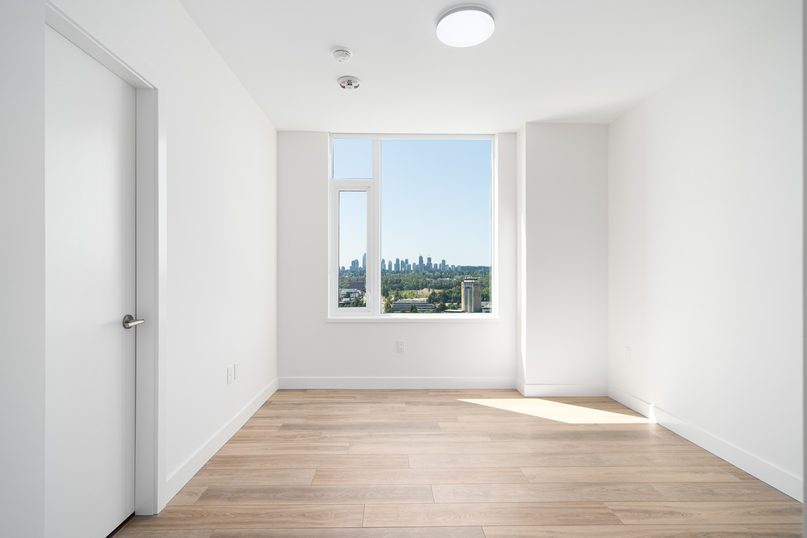 Empty, modern room with white walls, wood flooring, large window, and a view of a distant city skyline under a clear sky.